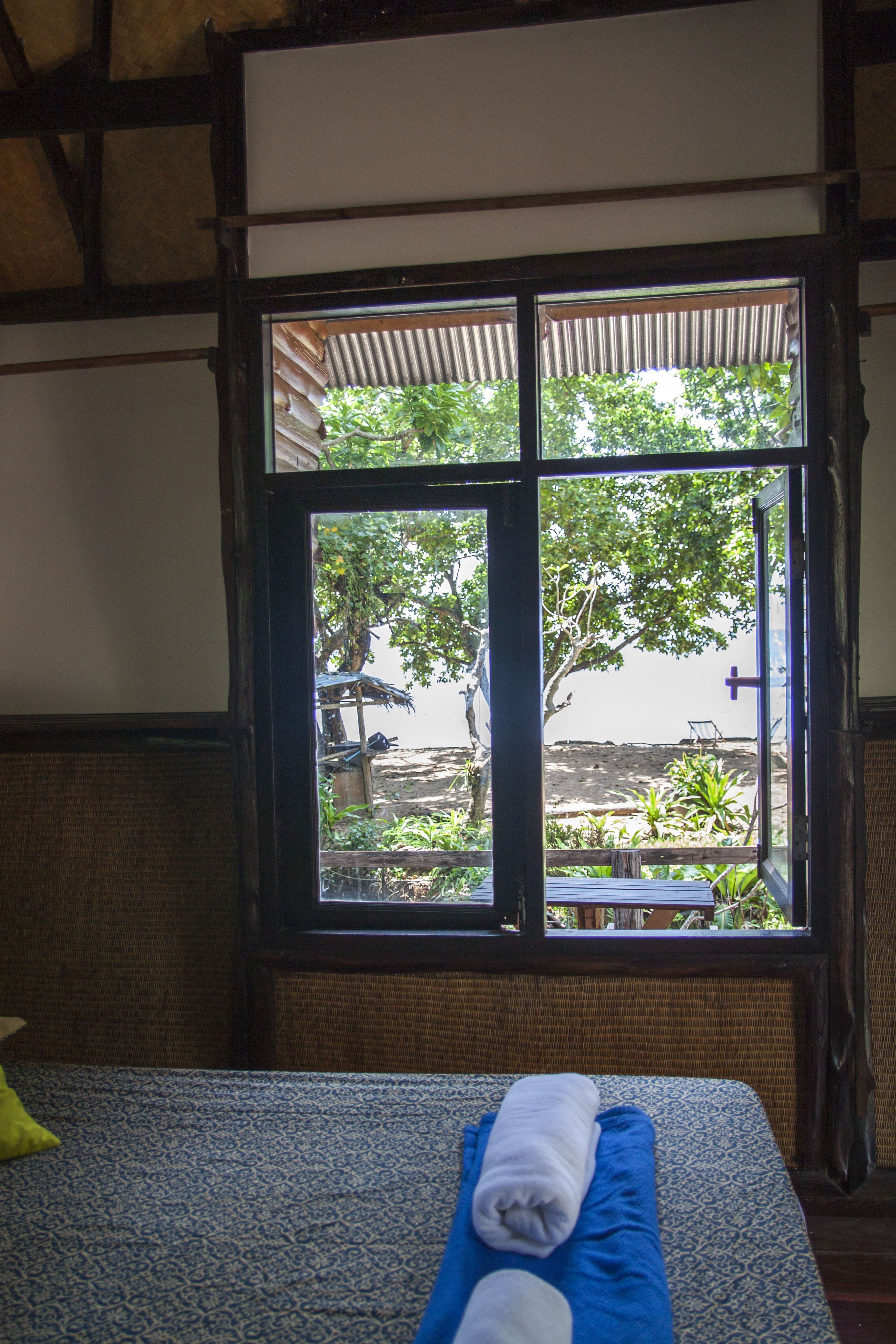 A room with a bed, a window with a view of greenery outside, and a wooden ceiling.