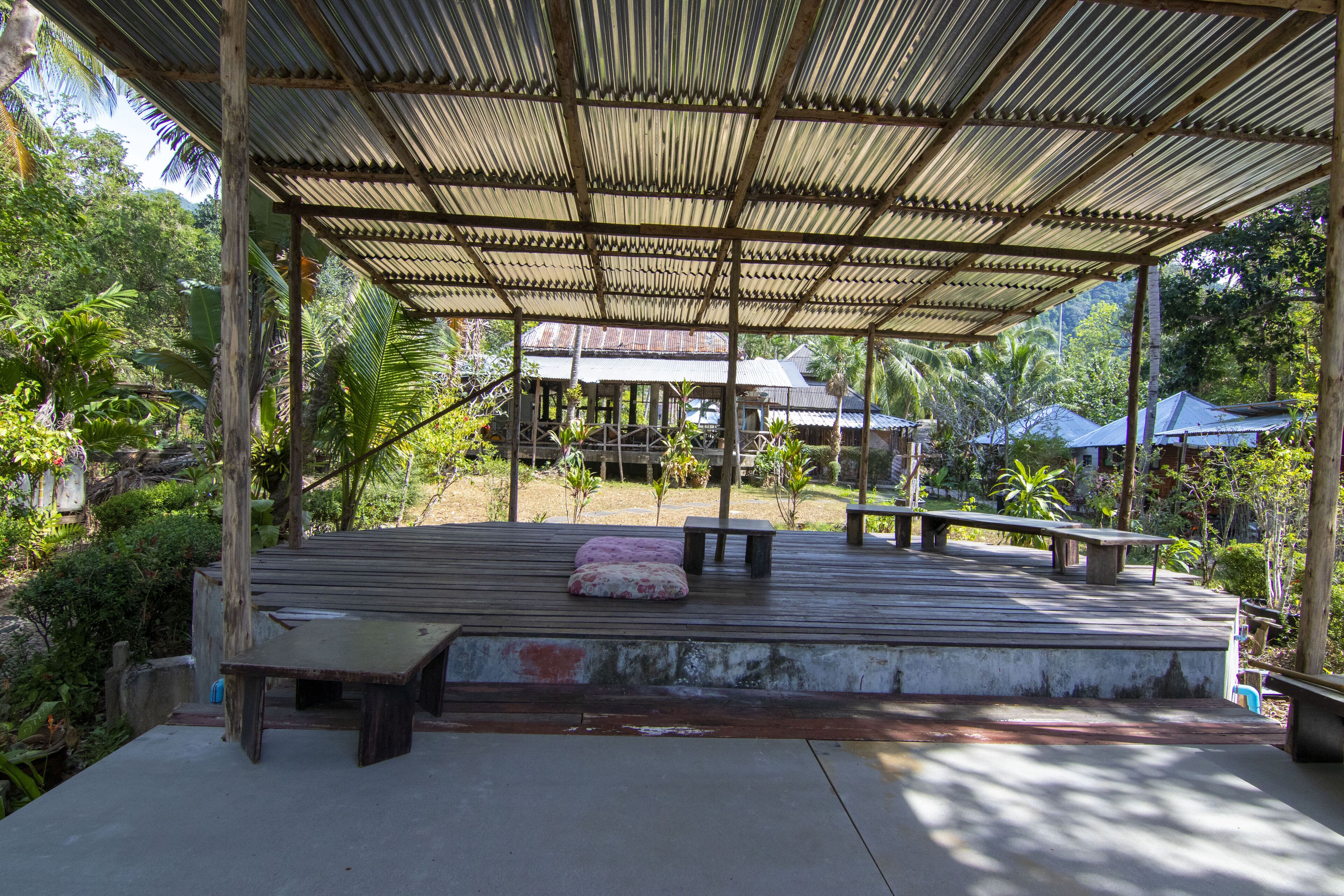 Un espacio de estar al aire libre cubierto, con bancos y una mesa, rodeado de abundante vegetación y una casa al fondo.