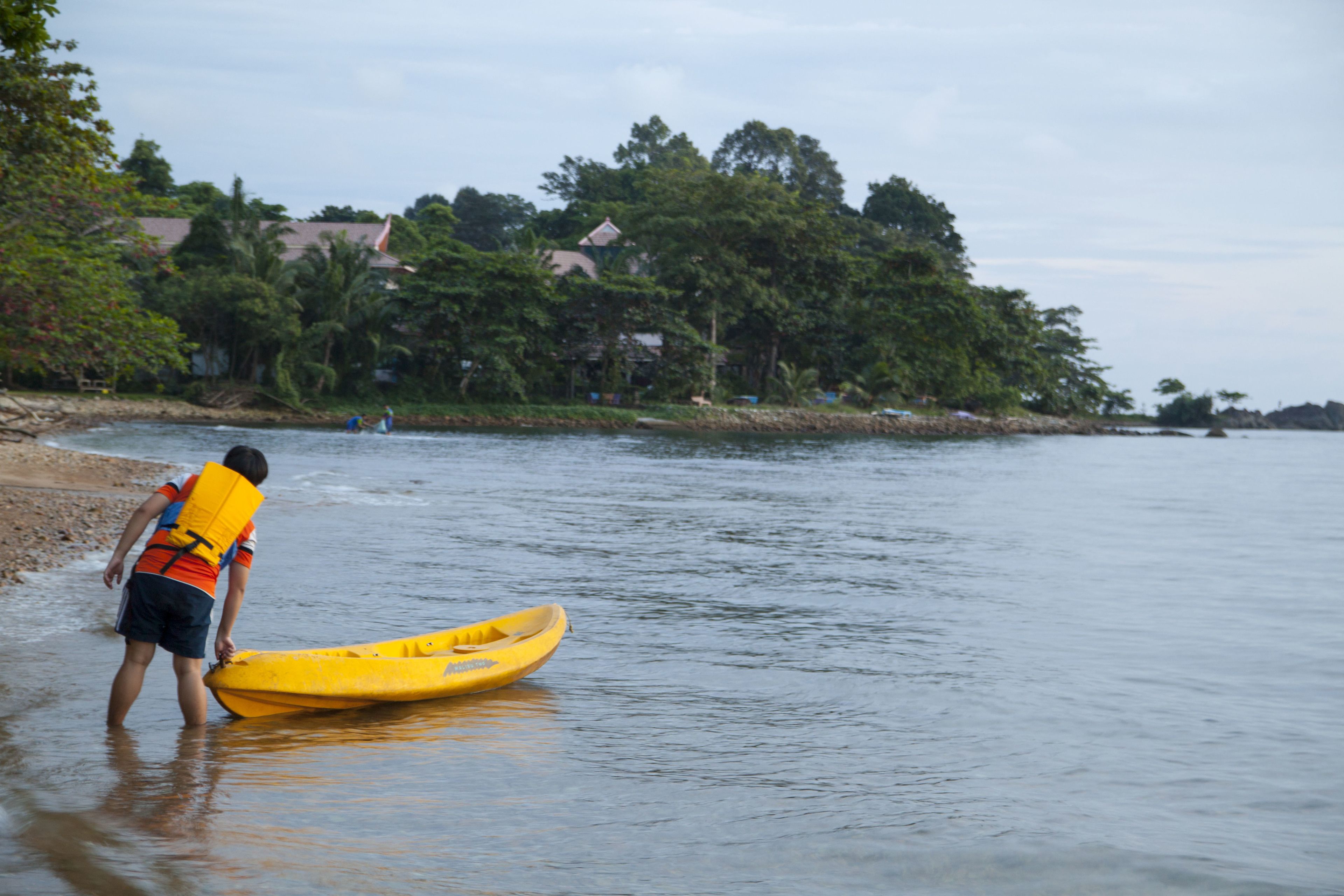 A person in a life jacket standing next to a yellow kayak on a calm body of water.