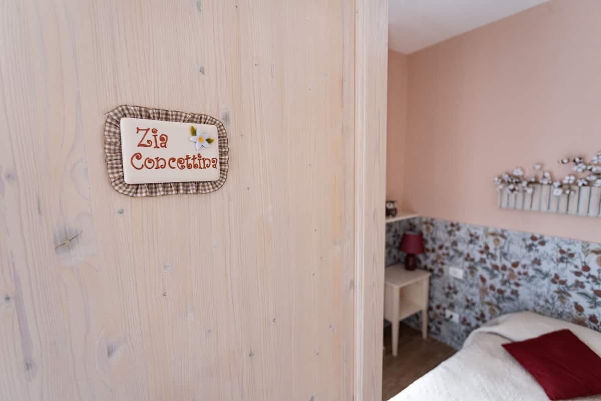 Ein Zimmer mit Holztür, einem Bett mit rotem Kissen, einem kleinen Tisch mit Lampe und blumigem Wandpapier.