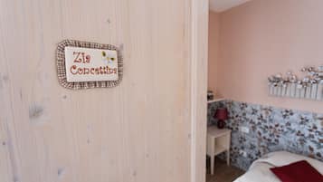 Una habitación con puerta de madera, una cama con almohada roja, una mesita con lámpara y papel tapiz floral en la pared.