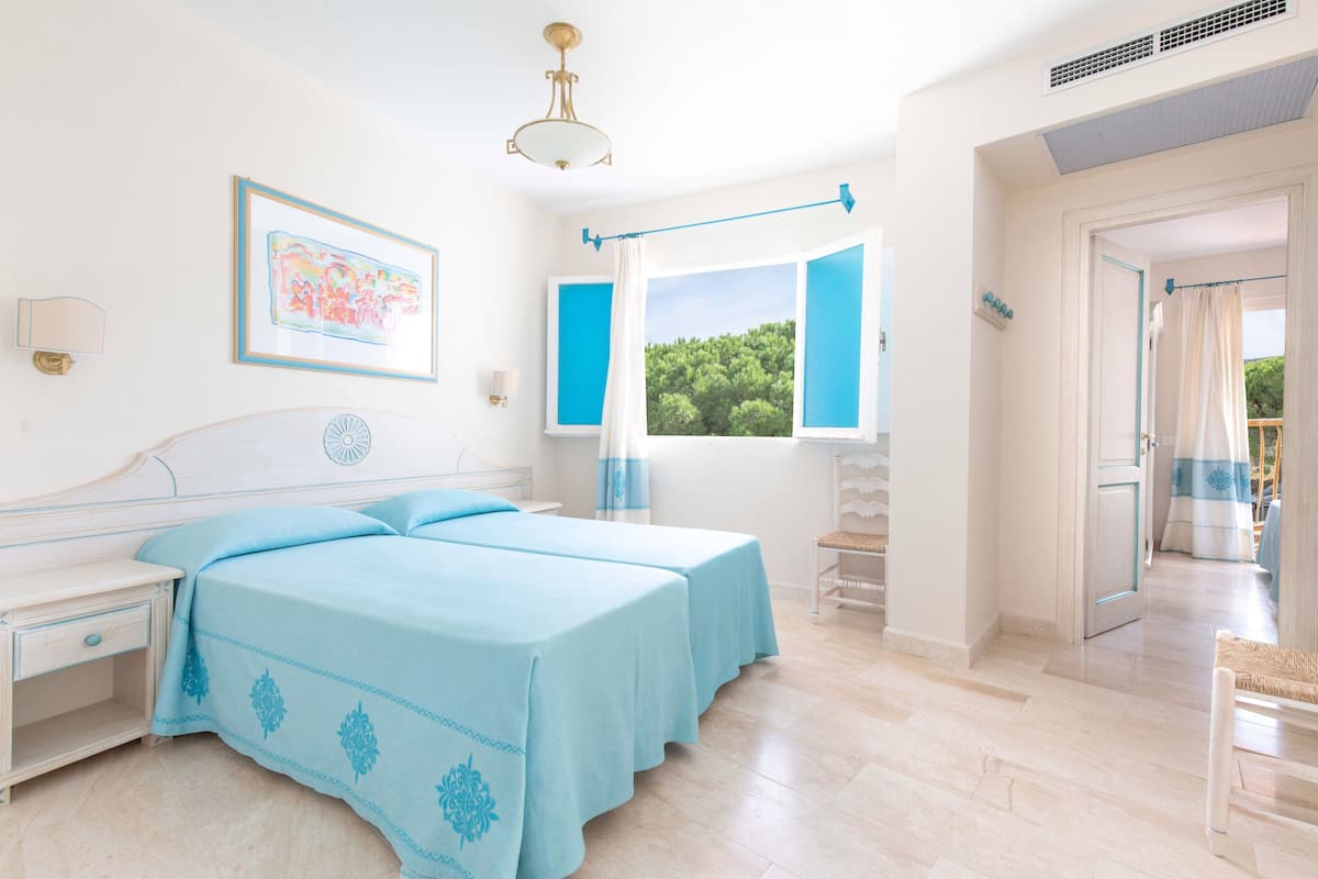A bedroom with two beds, a nightstand, a window with curtains, and a view of greenery outside.