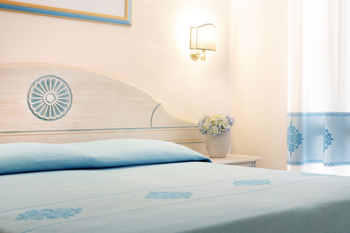 A neatly made bed with a headboard featuring a decorative design, a bedside table with a vase of flowers, and a wall-mounted light fixture.