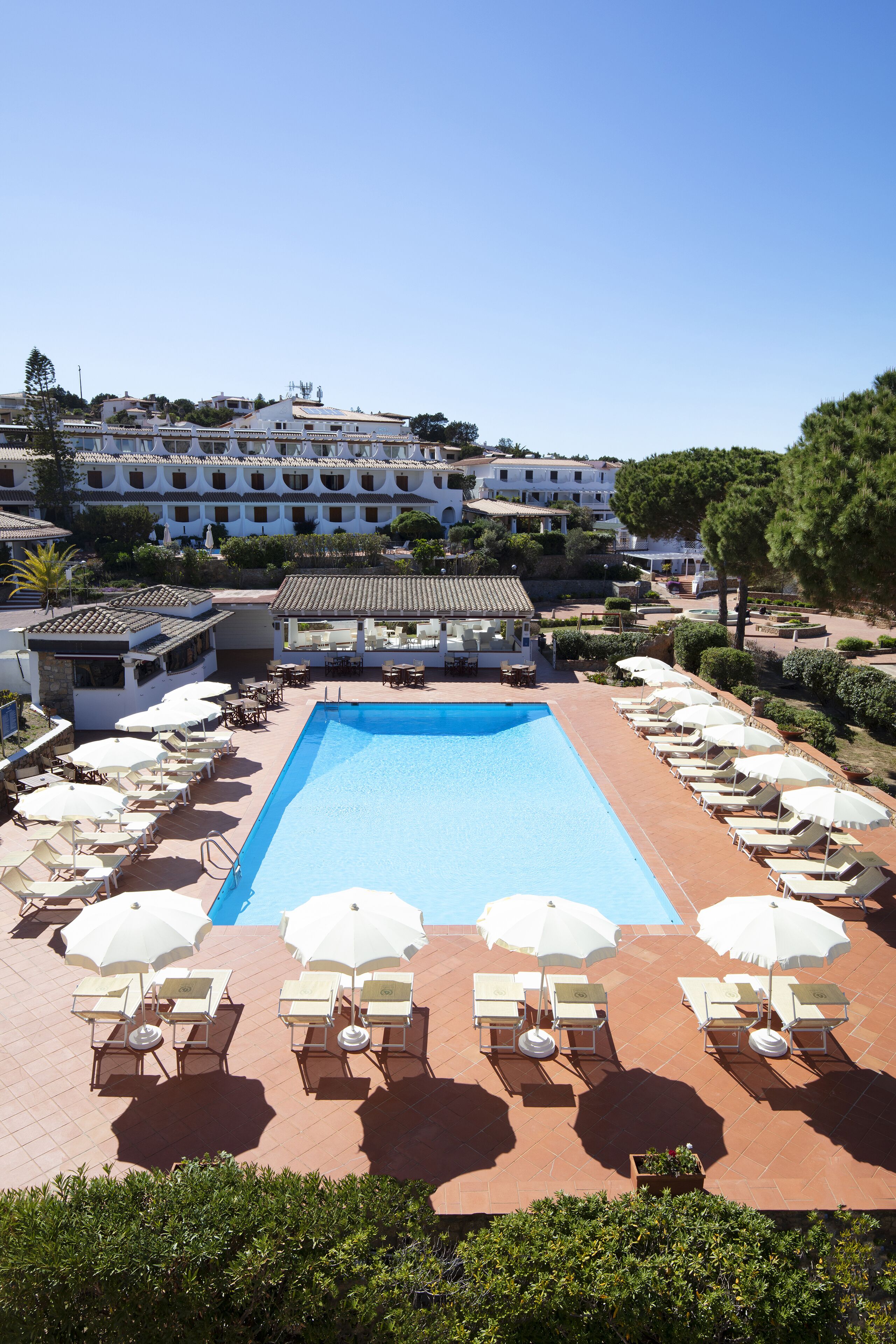 A swimming pool with sun loungers and umbrellas, surrounded by a building with multiple floors and balconies.