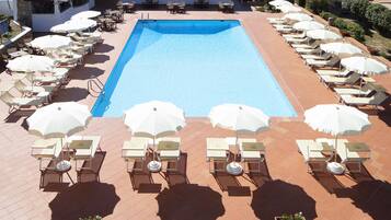 A swimming pool with sun loungers and umbrellas, surrounded by a building with multiple floors and balconies.