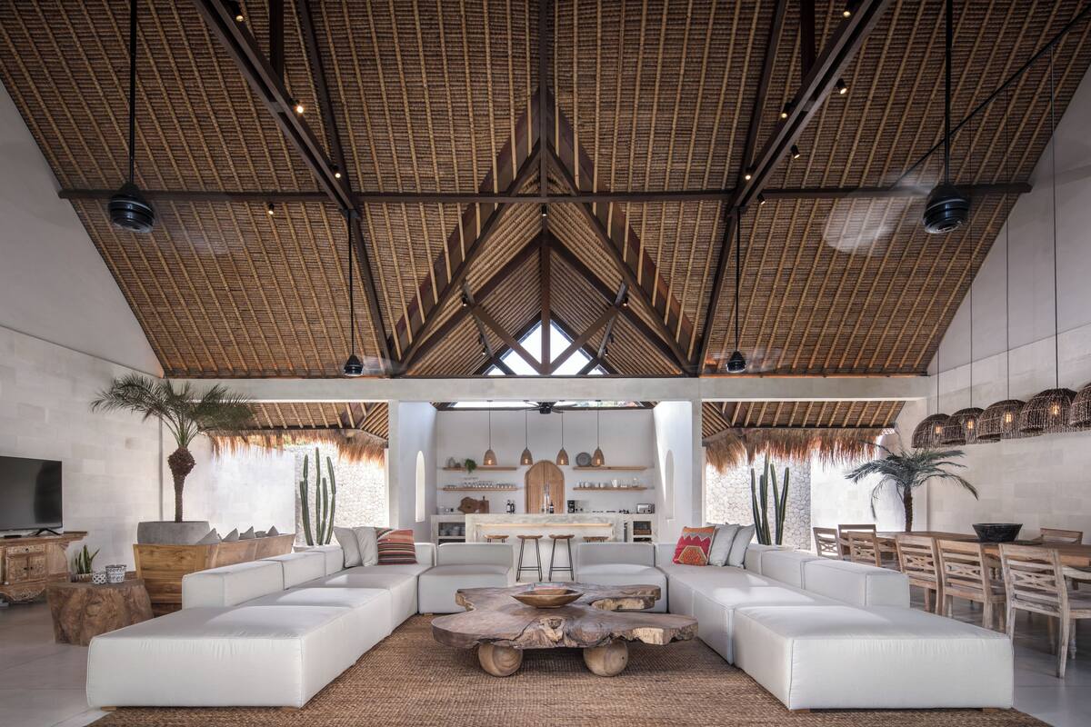 A spacious living area with a high ceiling and a thatched roof design, featuring a large L-shaped sectional sofa, a wooden coffee table, and a dining area with wooden chairs.