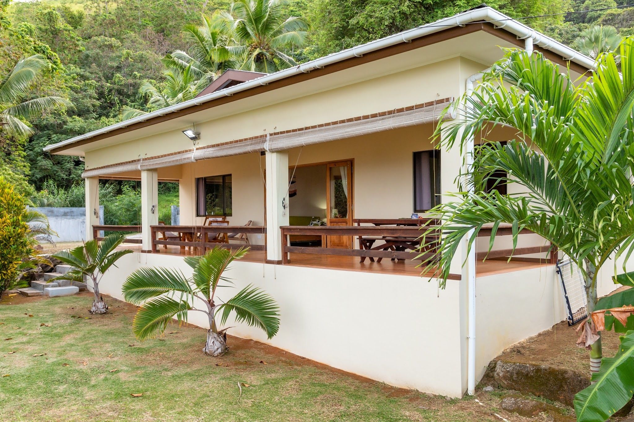 A two-story house with a covered patio area, surrounded by lush greenery and palm trees.