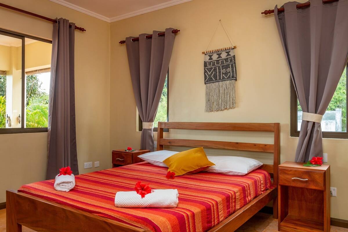 A hotel room with a wooden bed, grey curtains, a macrame wall hanging, and a red-striped bedspread.