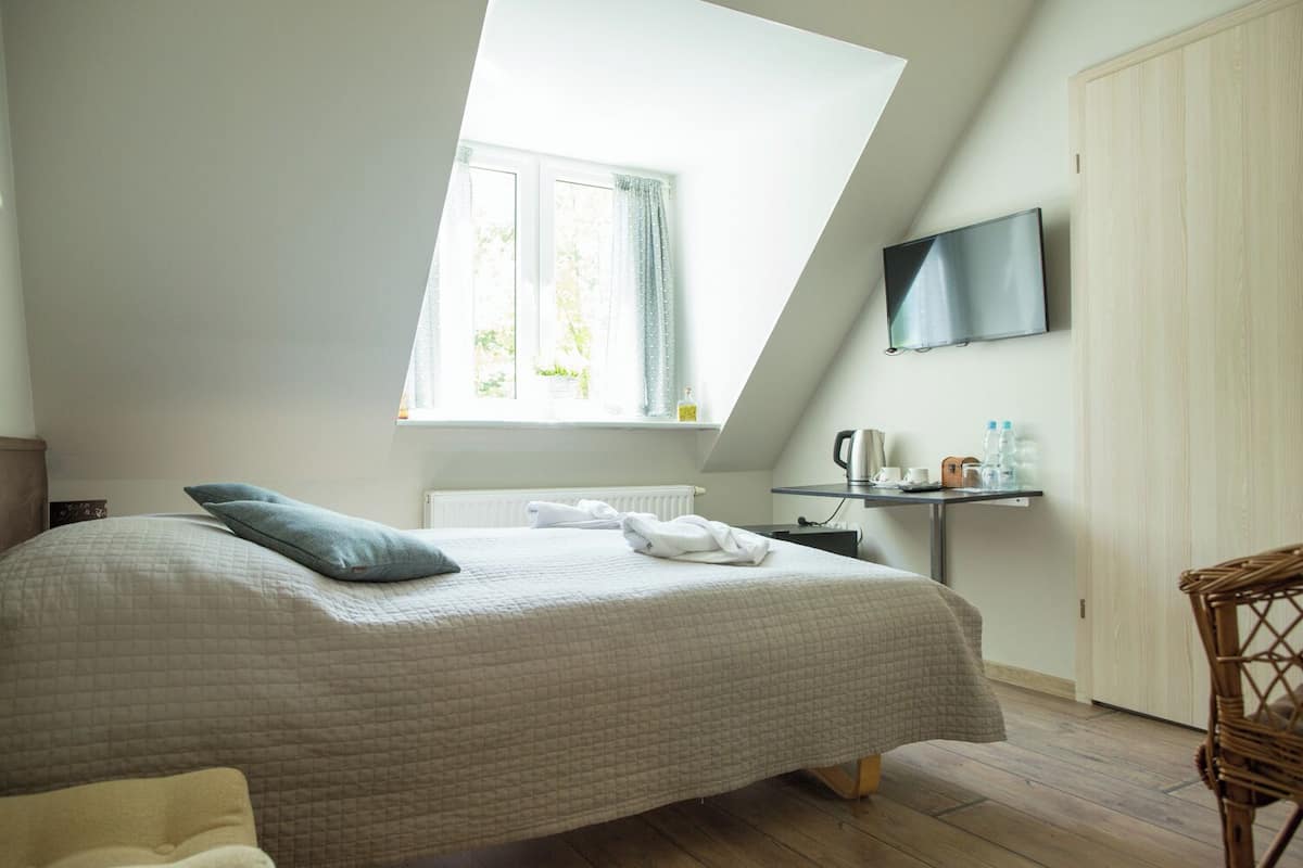 A bedroom with a sloped ceiling, a bed with a quilted bedspread, a window with curtains, a flat-screen TV mounted on the wall, a small table with a kettle and cups, and a wicker chair.