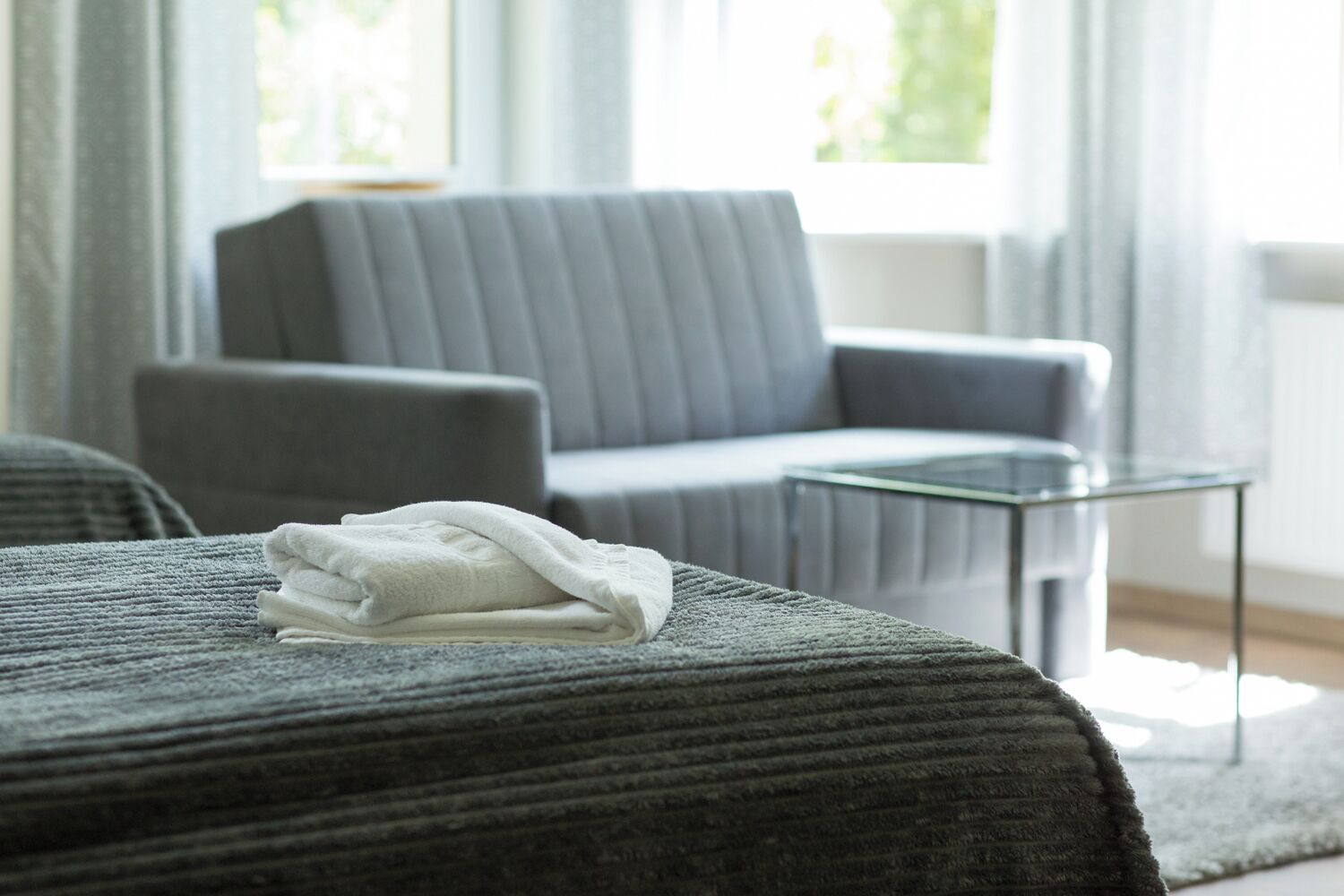 Une chambre d’hôtel moderne avec un canapé gris, une table basse en verre et une serviette blanche soigneusement pliée sur un lit gris.