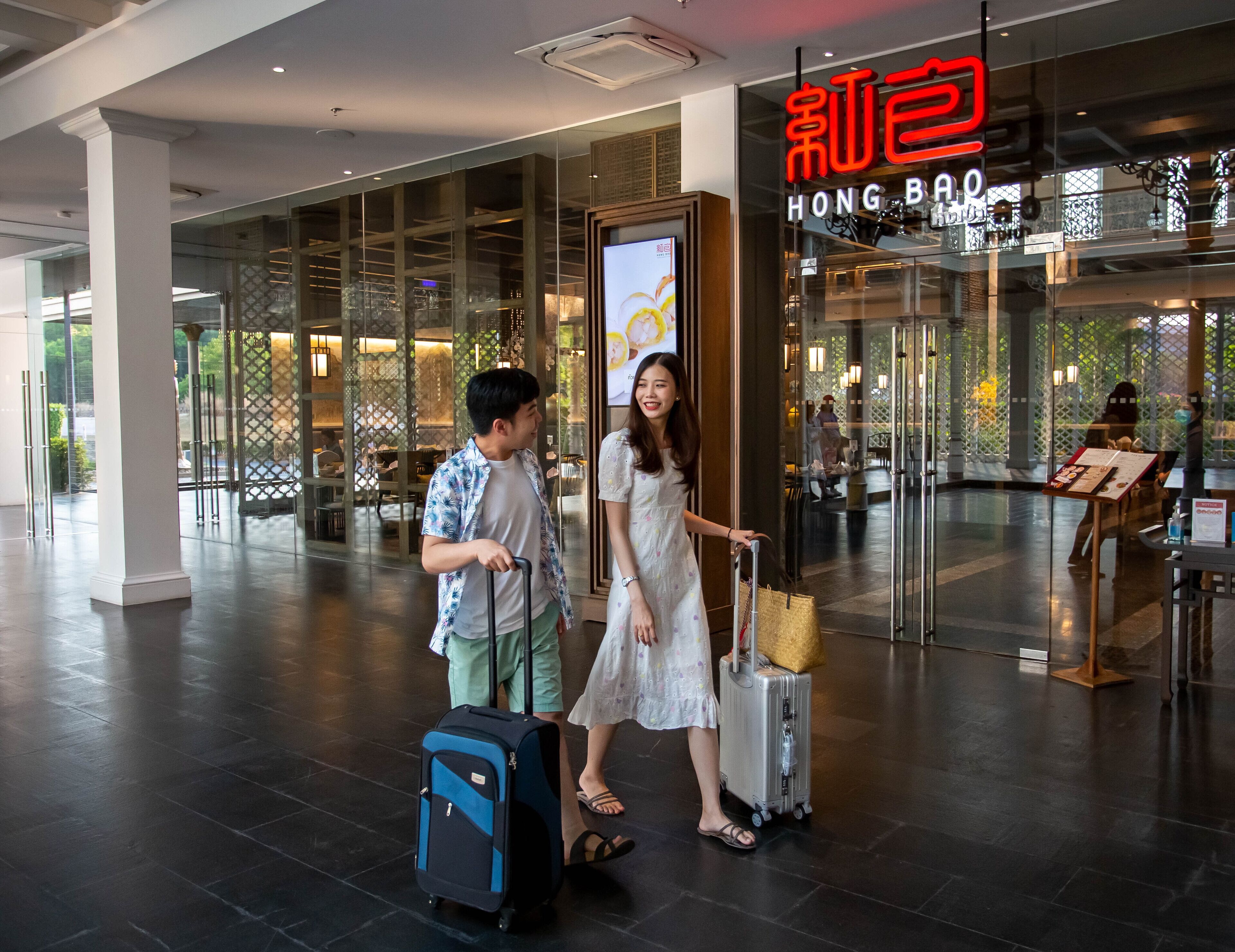 Un hall d’hôtel moderne avec une façade en verre, un espace restaurant bien visible et une enseigne indiquant « HONG BAO ».