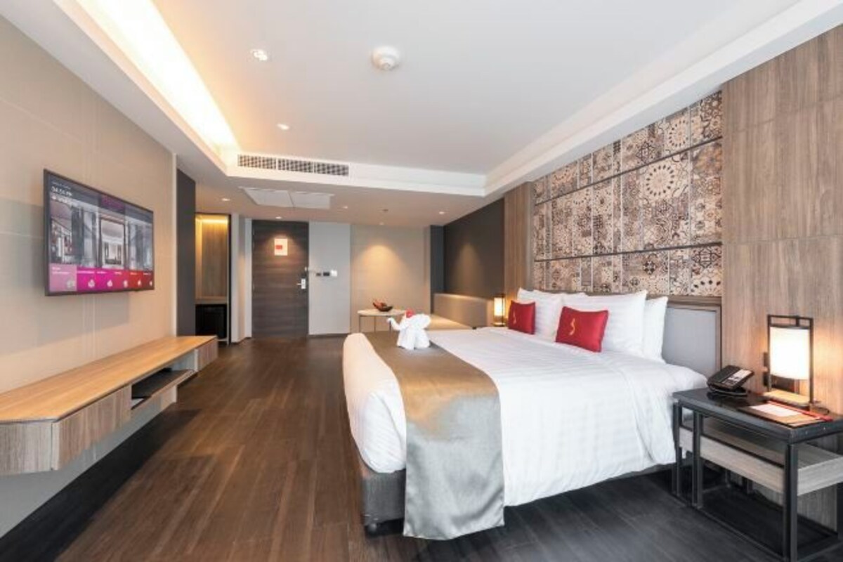 A modern hotel room with a large bed, a flat-screen TV, a desk with a lamp, and a patterned wall.
