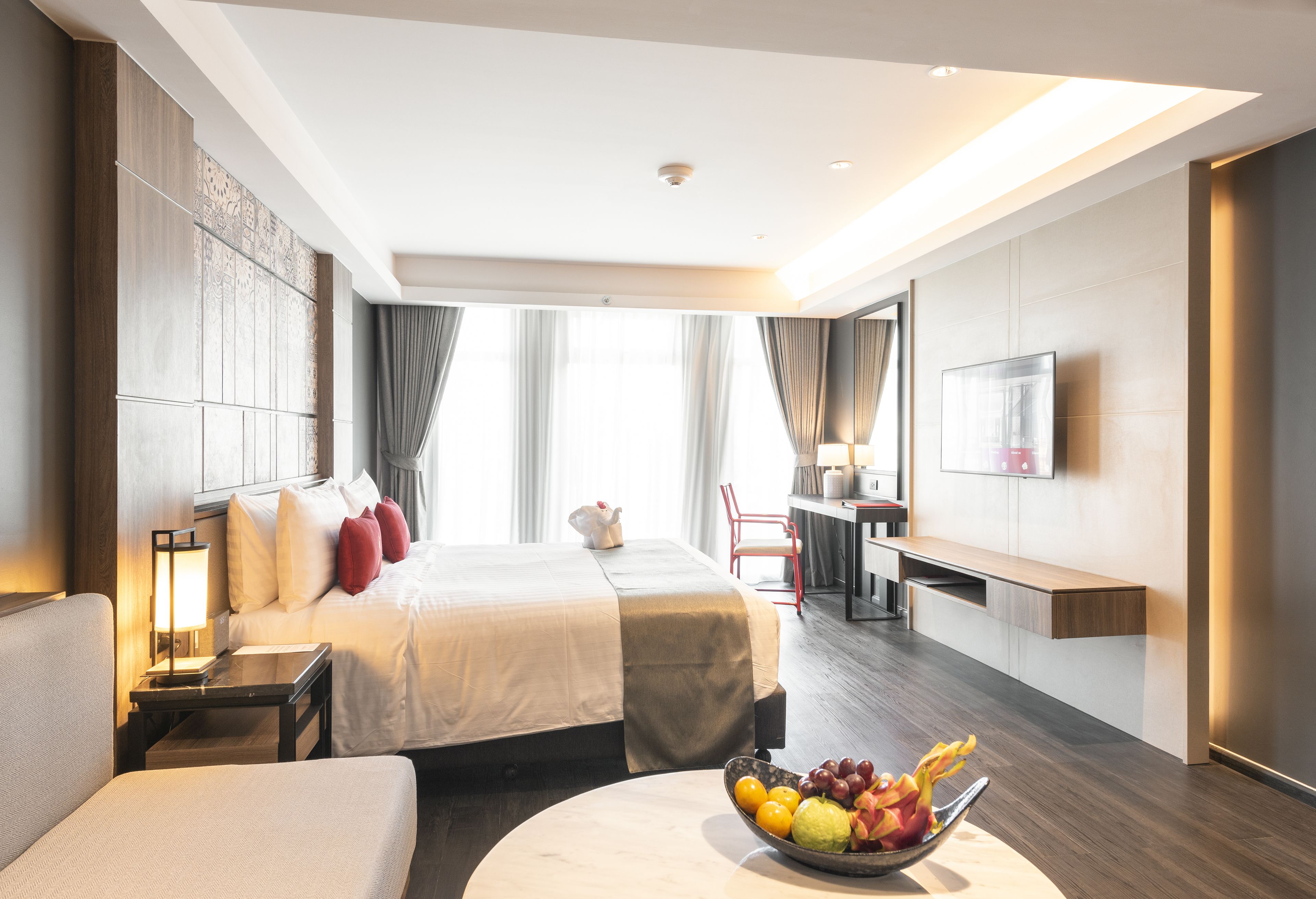 A modern hotel room with a large bed, bedside tables, a desk, a flat-screen TV, and a fruit bowl on a round table.