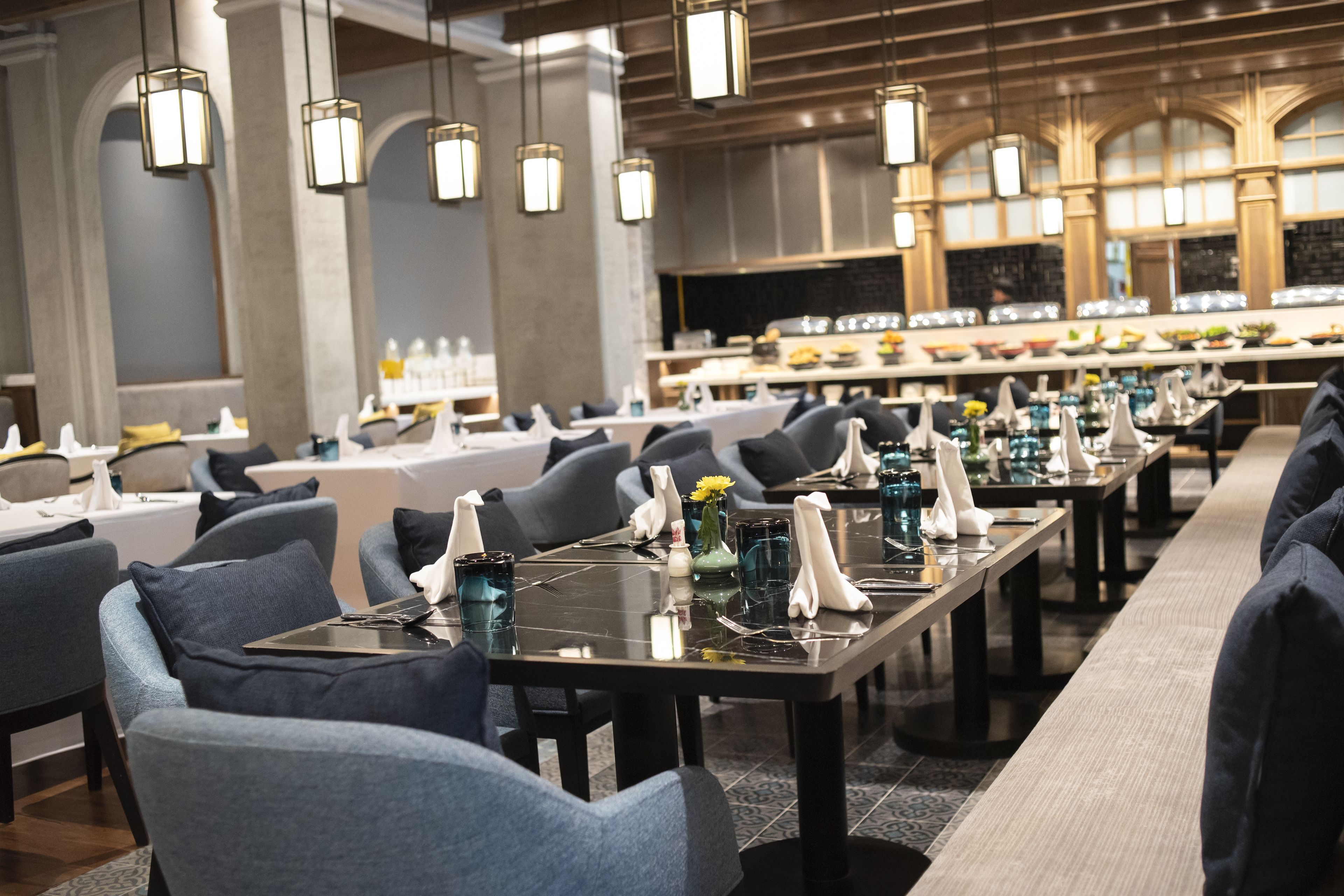 A modern restaurant with long tables, grey upholstered chairs, and pendant lighting.