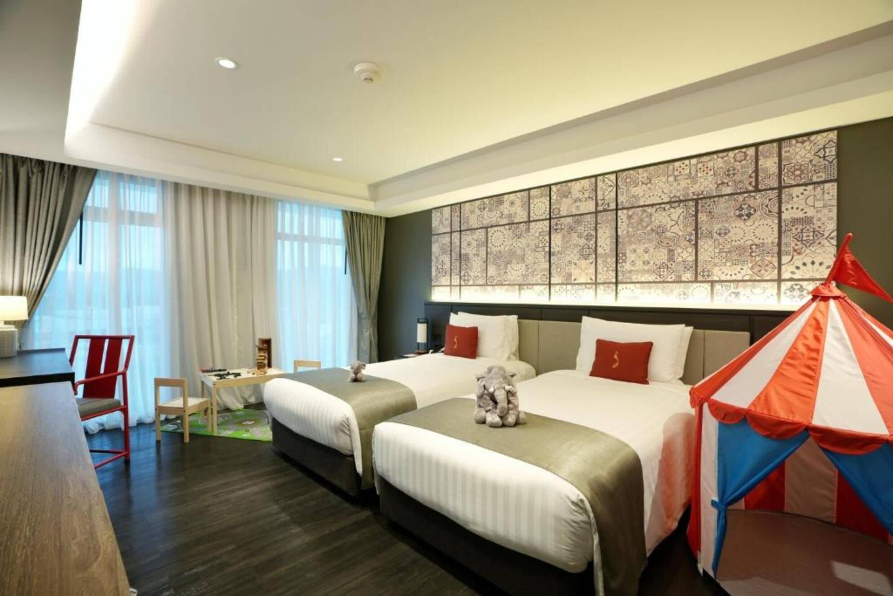 A hotel room with two beds, a desk, a chair, a small table, and a decorative wall panel.