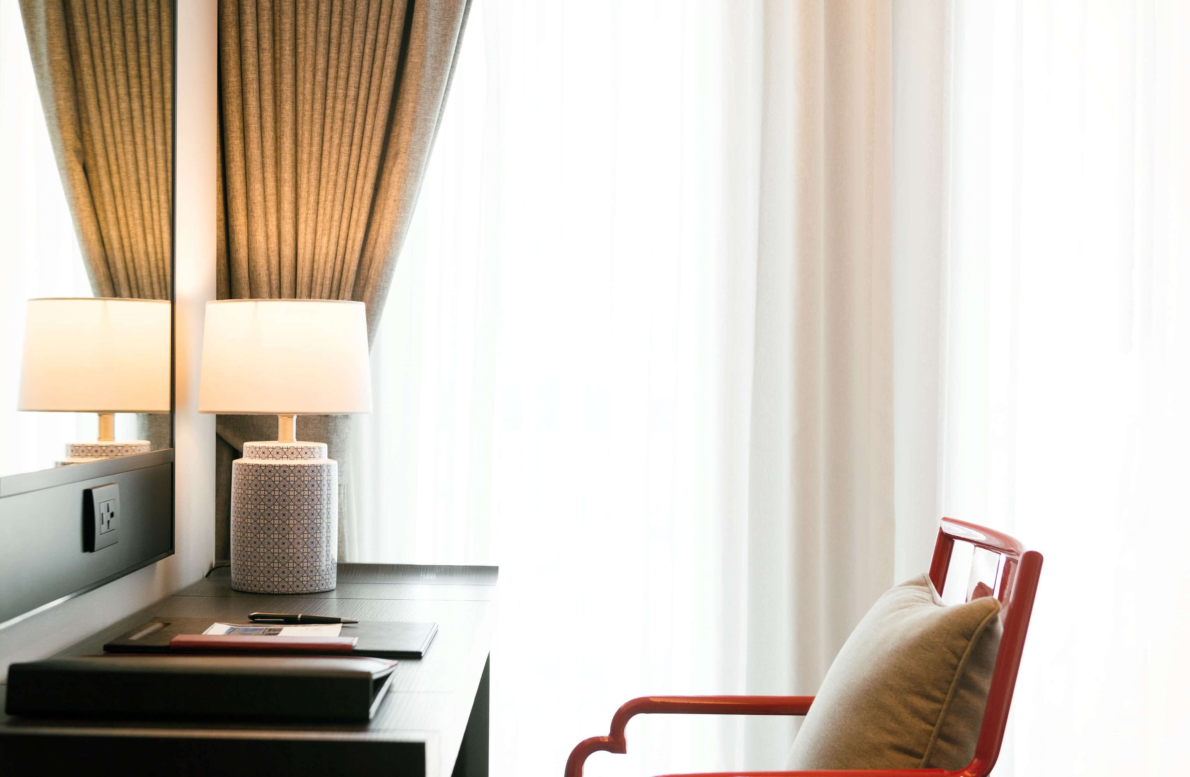 Una habitación de hotel con escritorio, dos lámparas, una silla roja y una ventana con cortinas.