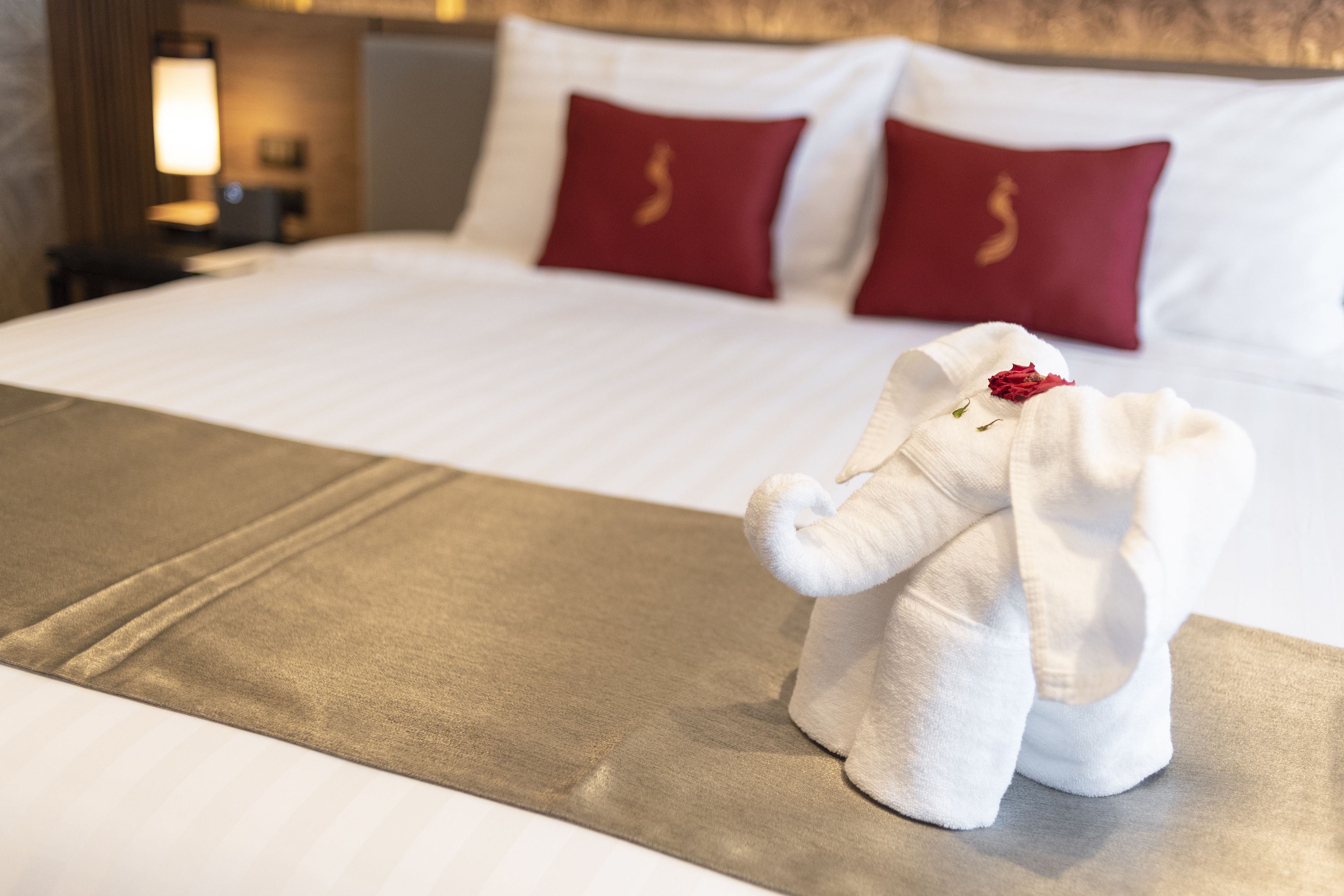 A neatly made bed with a golden bedspread, two red pillows with a gold emblem, and a white towel elephant with a flower on a table.
