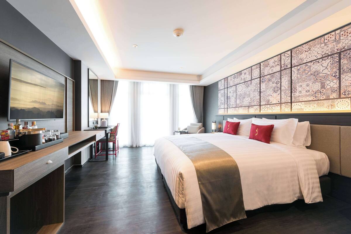 A modern hotel room with a large bed, a desk with a flat-screen TV, a dining area, and a decorative wall.