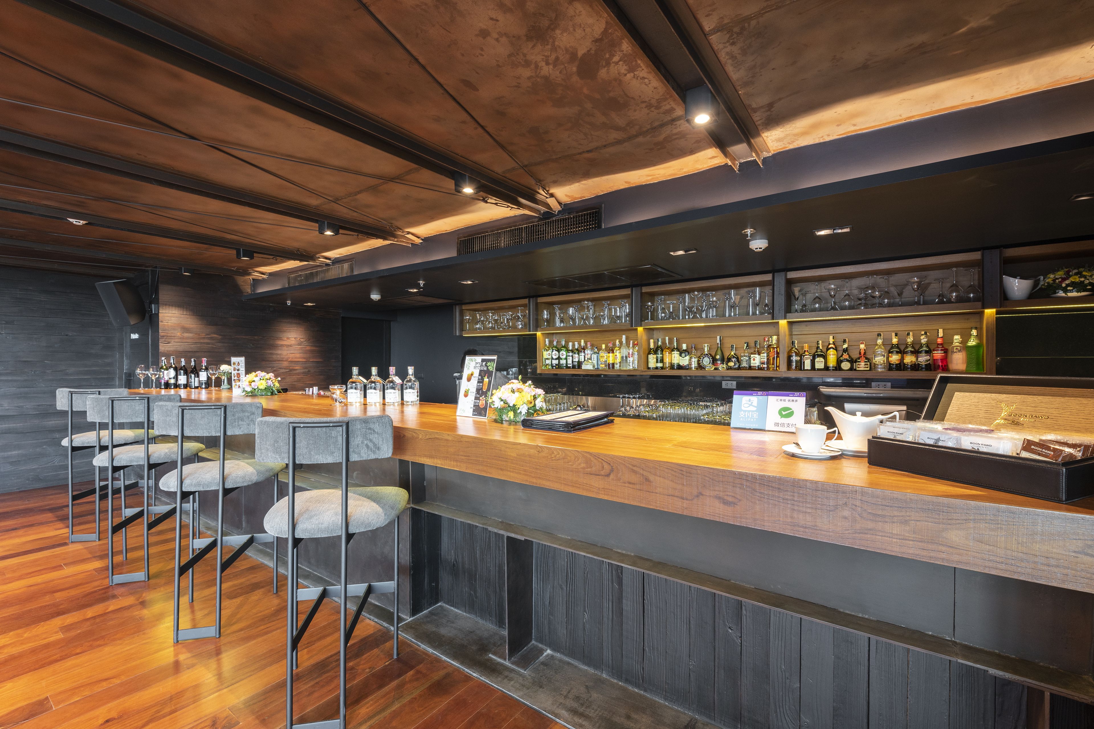 A bar area with a wooden counter, bar stools, shelves with bottles, glasses, and a menu board.