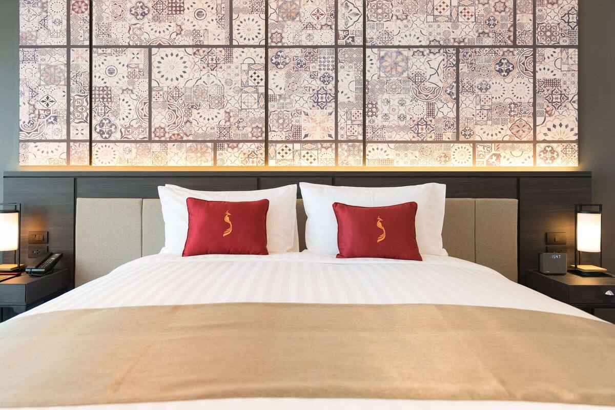 A hotel room with a large bed, two red pillows, a beige throw blanket, and a patterned wall above the bed.