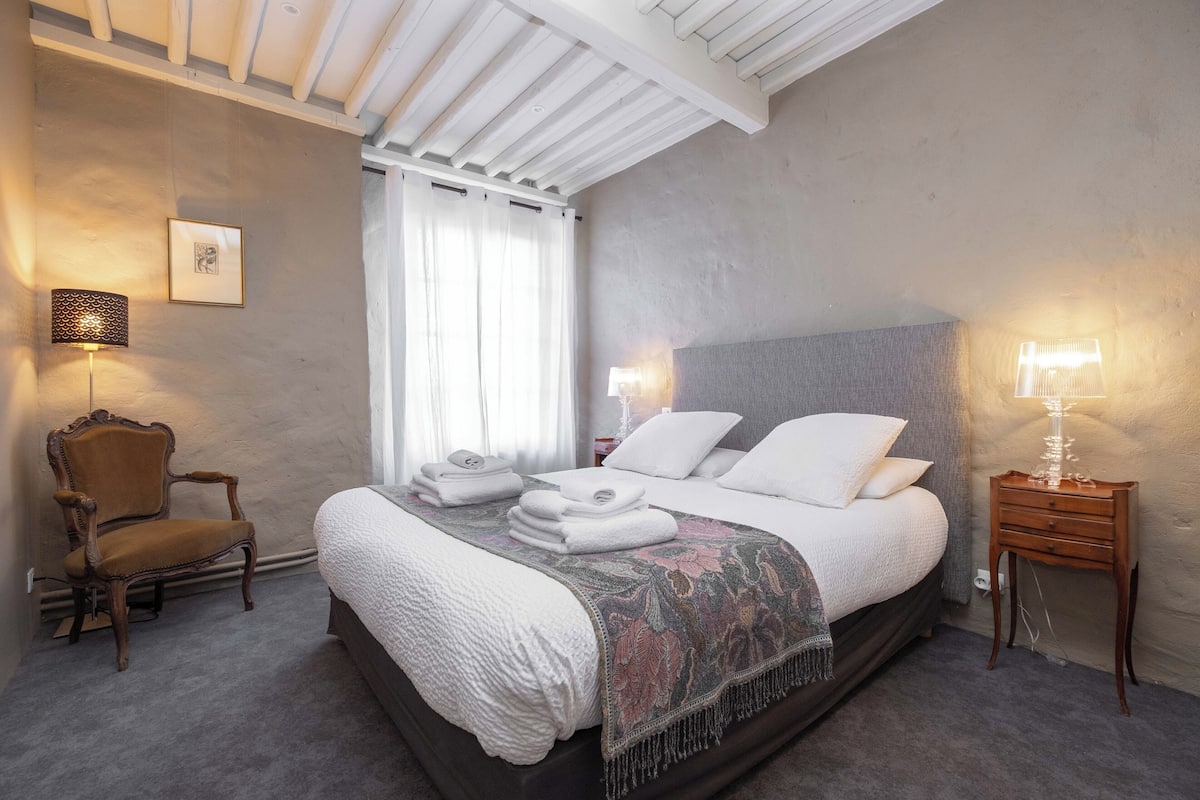 A bedroom with a large bed, two bedside lamps, a chair, and a window with curtains.