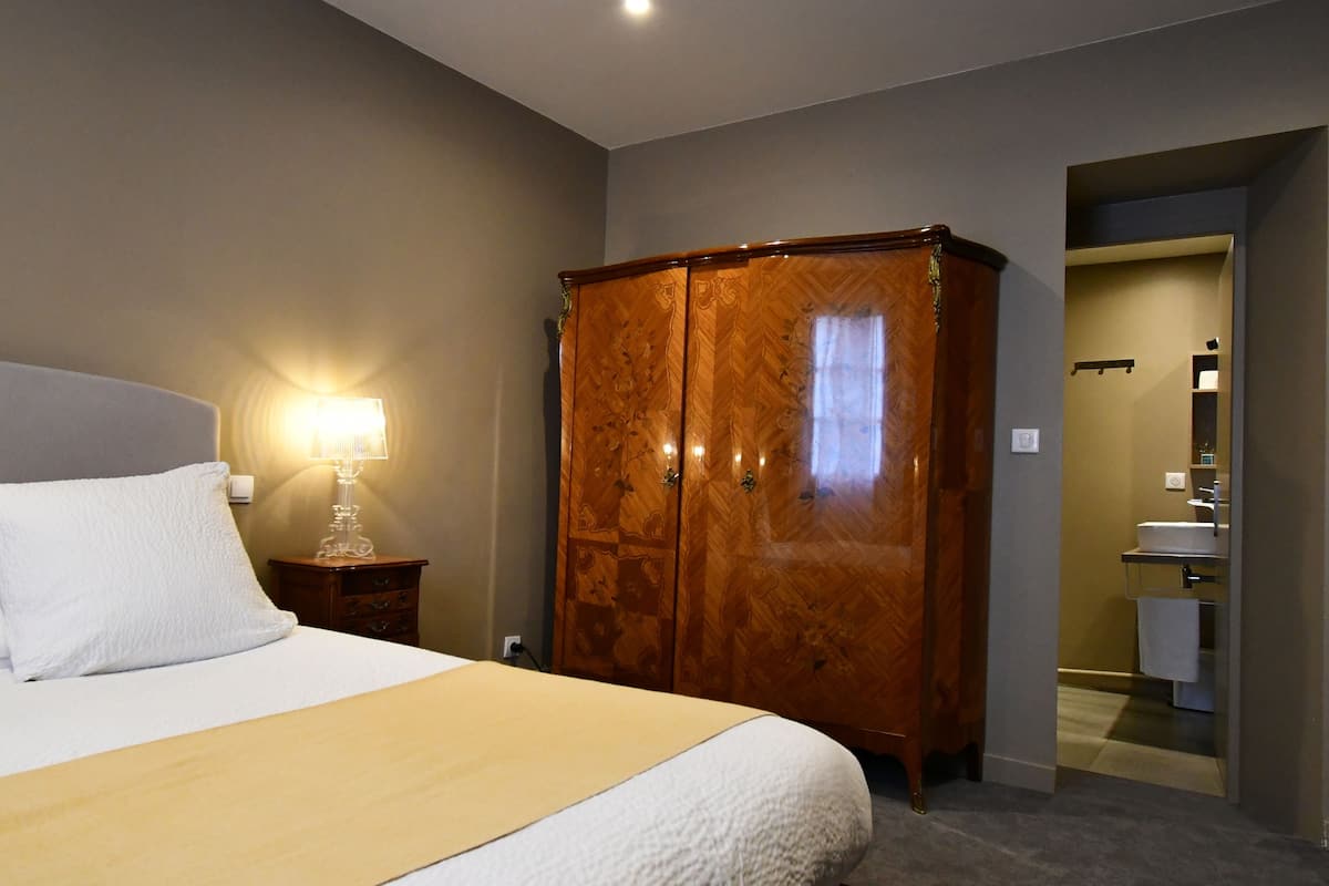 A hotel room with a bed, a wooden wardrobe, a nightstand with a lamp, and a bathroom visible through an open door.