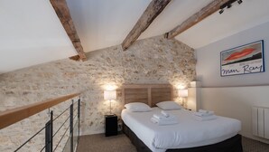 Un dormitorio con pared de piedra, viga de madera en el techo, cama con ropa blanca y una obra de arte enmarcada en la pared.