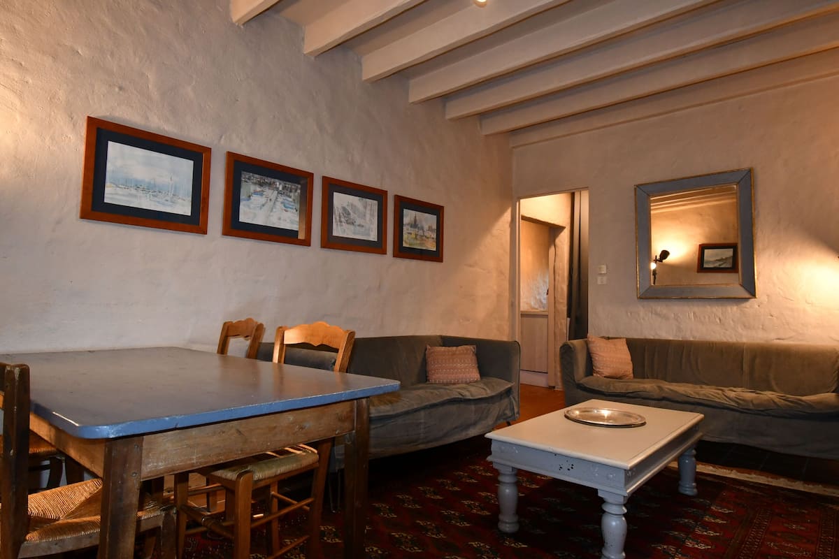 A room with a dining table, two wooden chairs, a sofa, and a small white table. There are four framed pictures on the wall.
