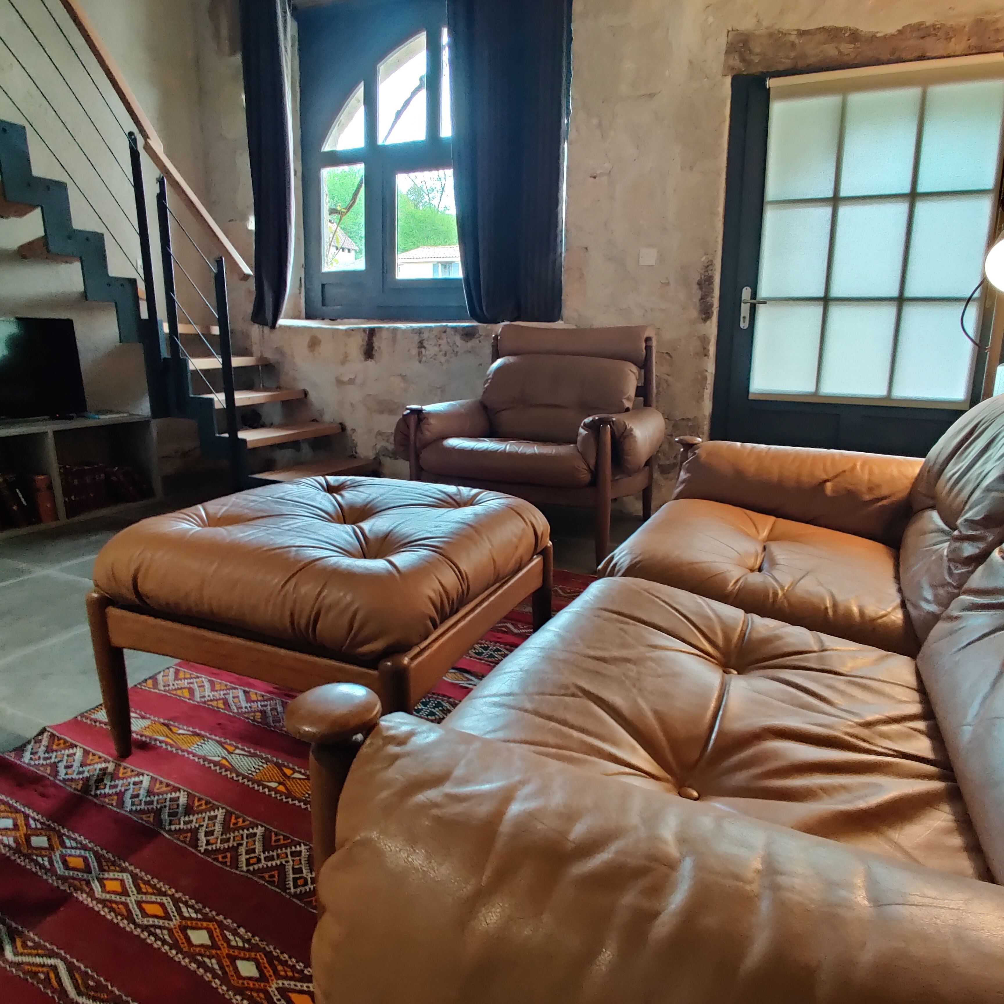 A living room with a sofa, armchair, and ottoman, a staircase, and a window with curtains.