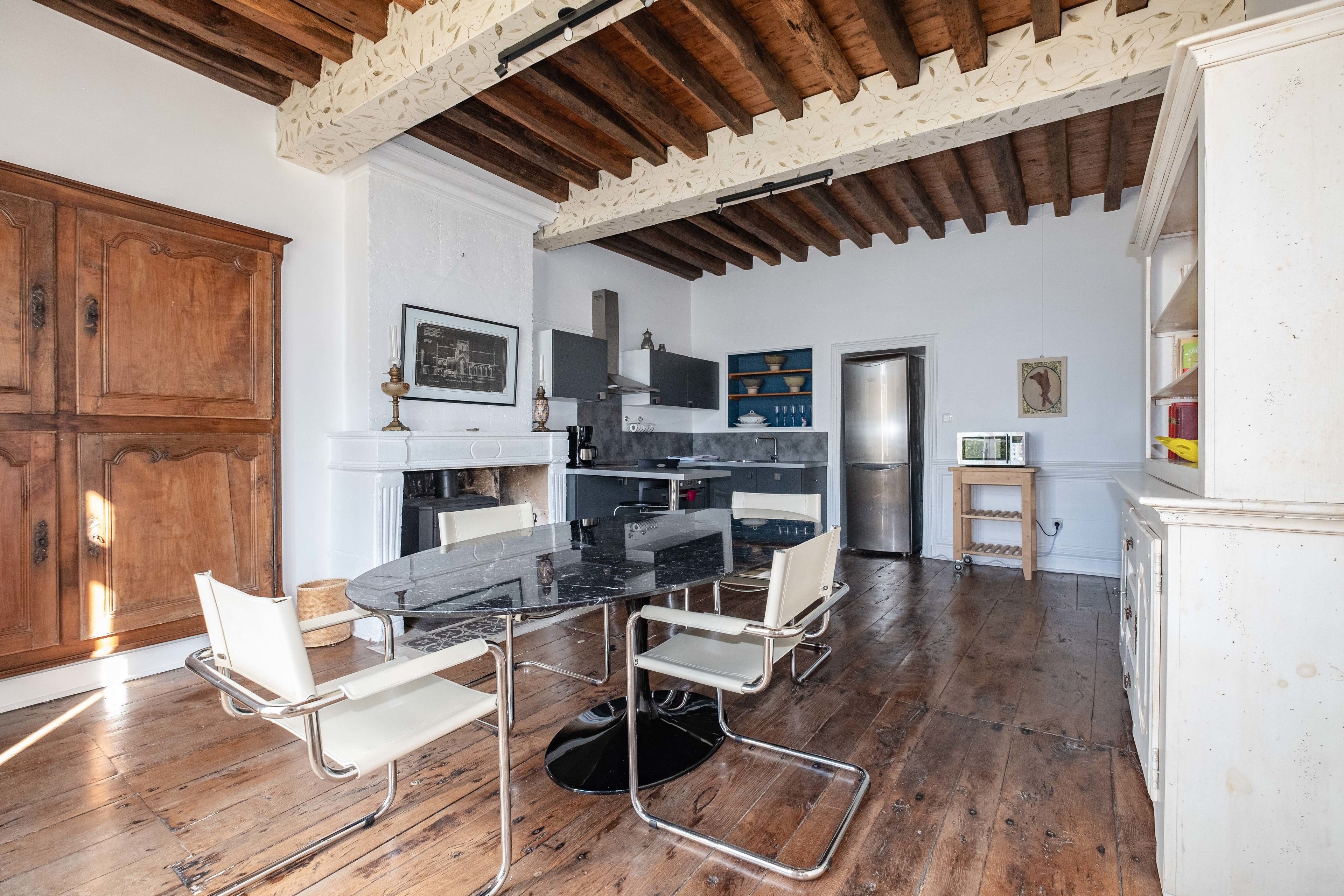 Comedor con una mesa de mármol negro y sillas blancas, piso de madera y techo rústico con vigas a la vista.