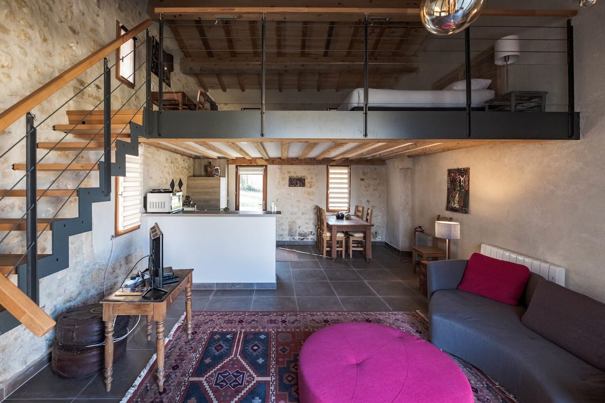 A spacious loft with a high ceiling, exposed wooden beams, a central island, a dining area, and a cozy seating area with a vibrant ottoman.