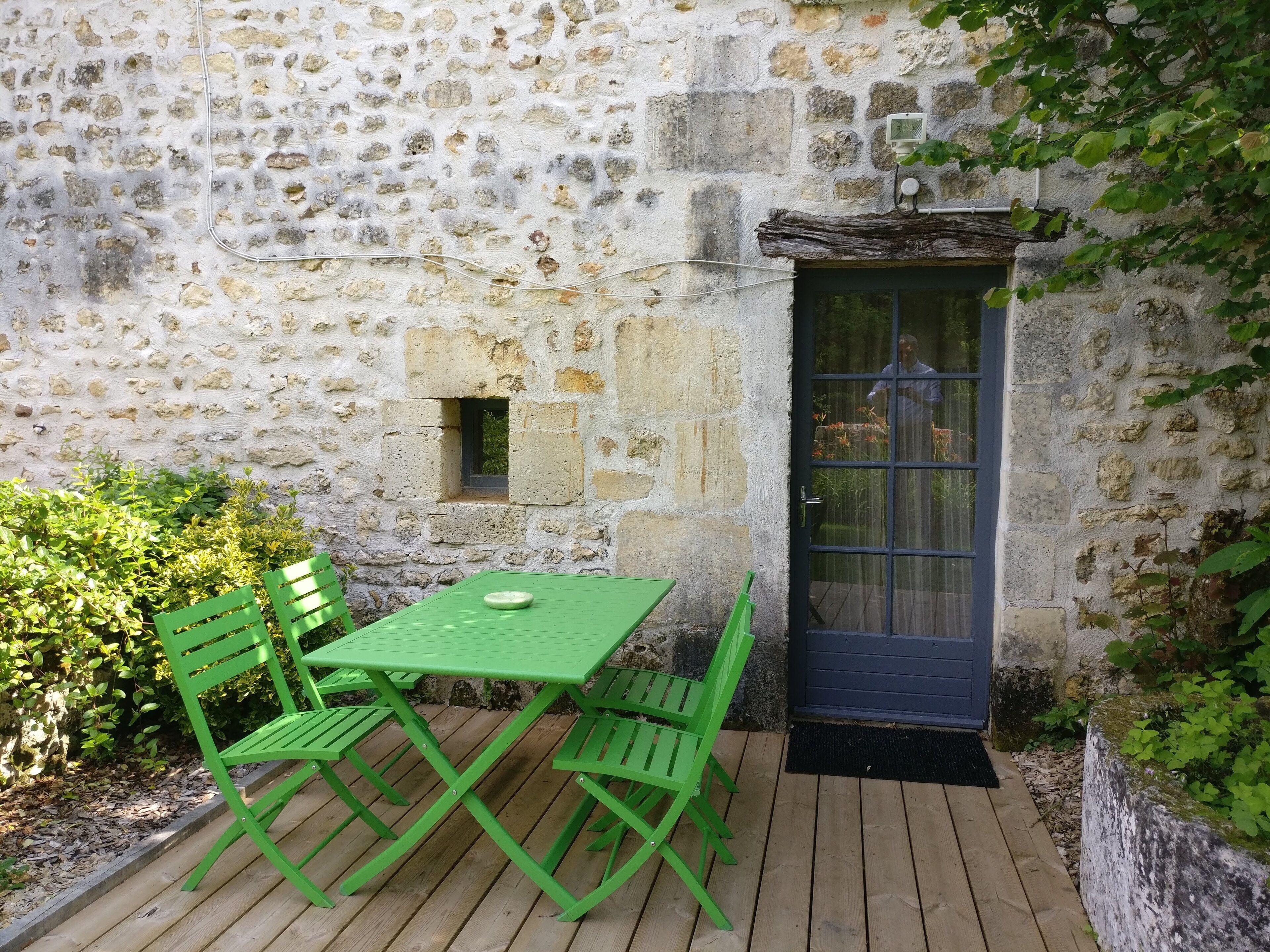 Een gebouw van steen met een houten terras, een groene tafel en bijpassende stoelen.