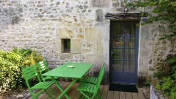 Een gebouw van steen met een houten terras, een groene tafel en bijpassende stoelen.