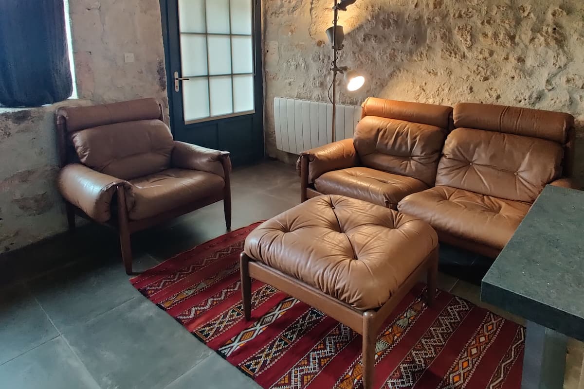 A rustic room with a stone wall, a wooden ceiling, a leather armchair, a sofa, a patterned rug, and a floor lamp.