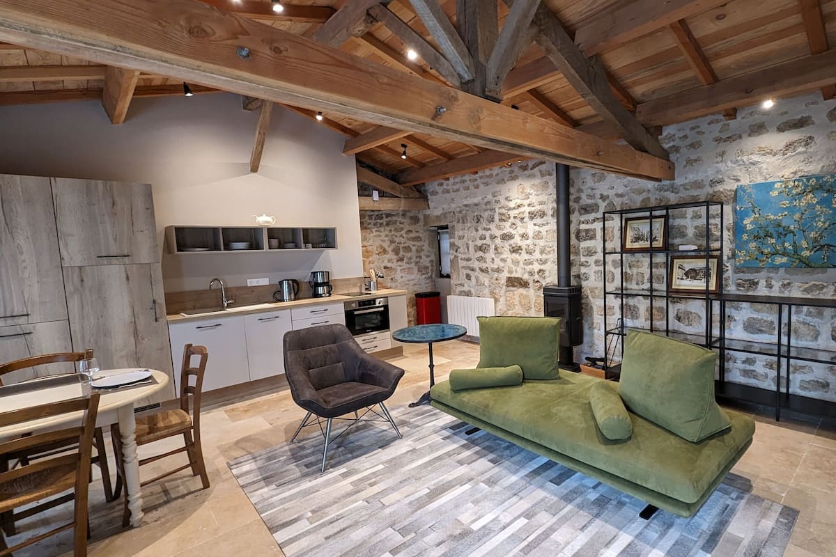 A cozy living room with a green sofa, a wooden dining table, and a kitchen area with stone walls and a fireplace.