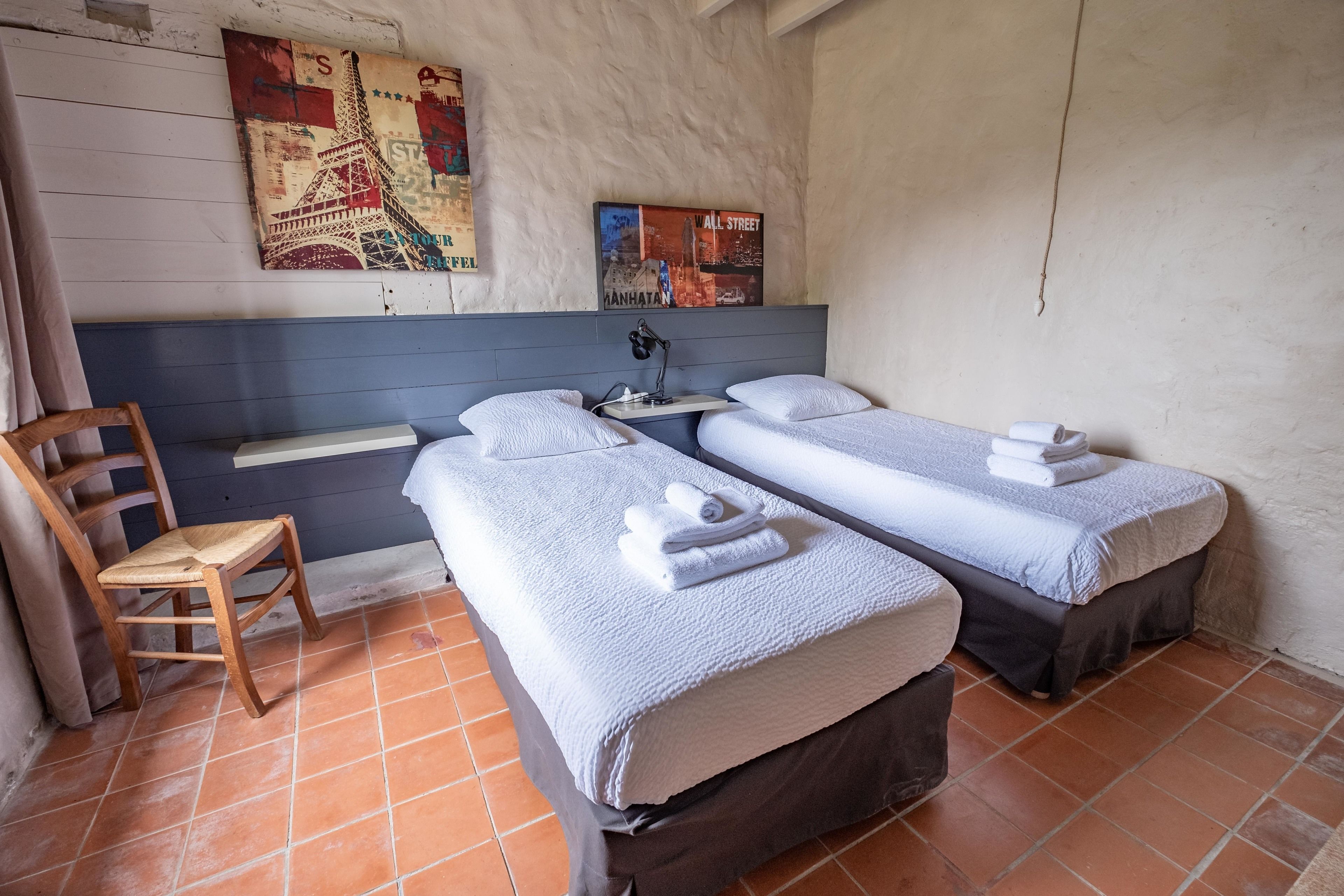 Una habitación con dos camas individuales, una silla, un escritorio con una lámpara y obras de arte en la pared.