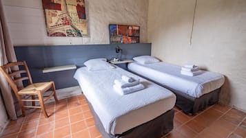 Una habitación con dos camas individuales, una silla, un escritorio con una lámpara y obras de arte en la pared.