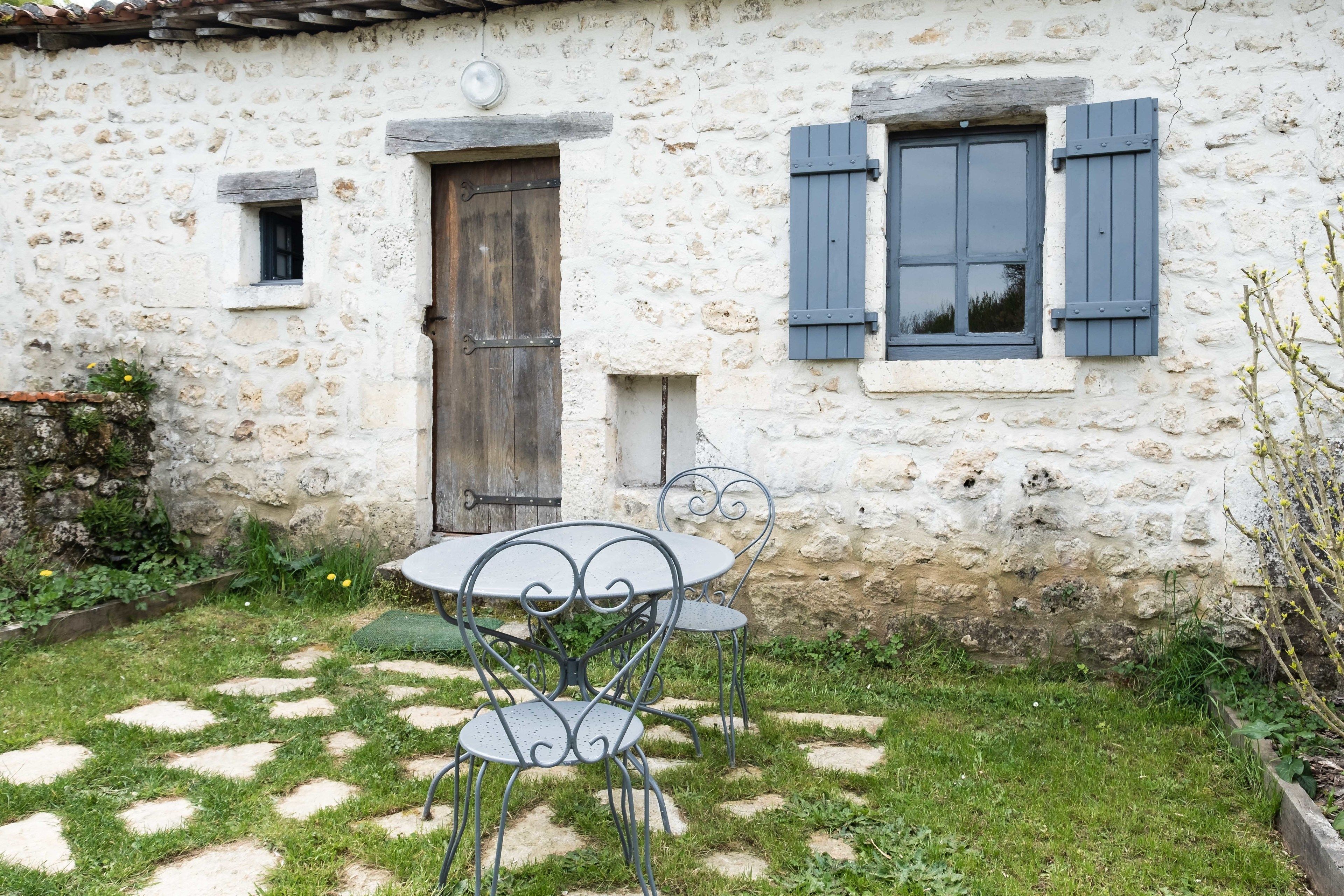 Un edificio rústico de piedra con puerta y contraventanas de madera, una ventana pequeña y un jardín con piedras para caminar y sillas de metal.