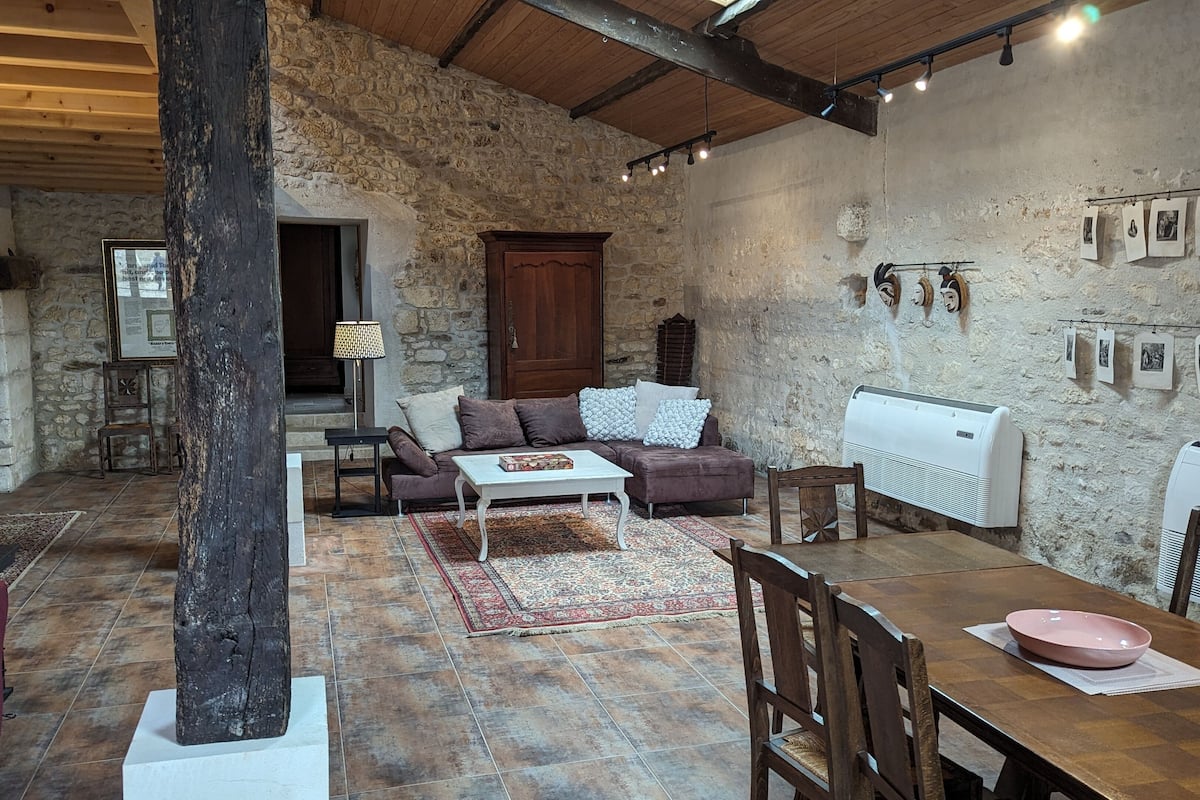 A rustic living room with exposed stone walls, a wooden ceiling, and a fireplace. There is a sofa, a coffee table, and a dining table with chairs.