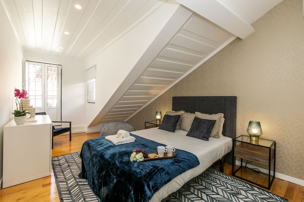 Ein Schlafzimmer mit geneigter Decke, einem Bett mit dunklem Kopfteil, Nachttischen mit Lampen, einem Schreibtisch mit Stuhl und einem Fenster mit Blick.