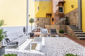 Un espace extérieur aménagé avec des meubles blancs, un mur en pierre et un bâtiment jaune en arrière-plan.