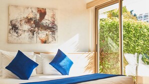 A hotel room with a bed, white bedding, blue pillows, a blue throw blanket, a painting on the wall, and a view of greenery through a window. - Mabrouk Hotel And Suites (Agadir)