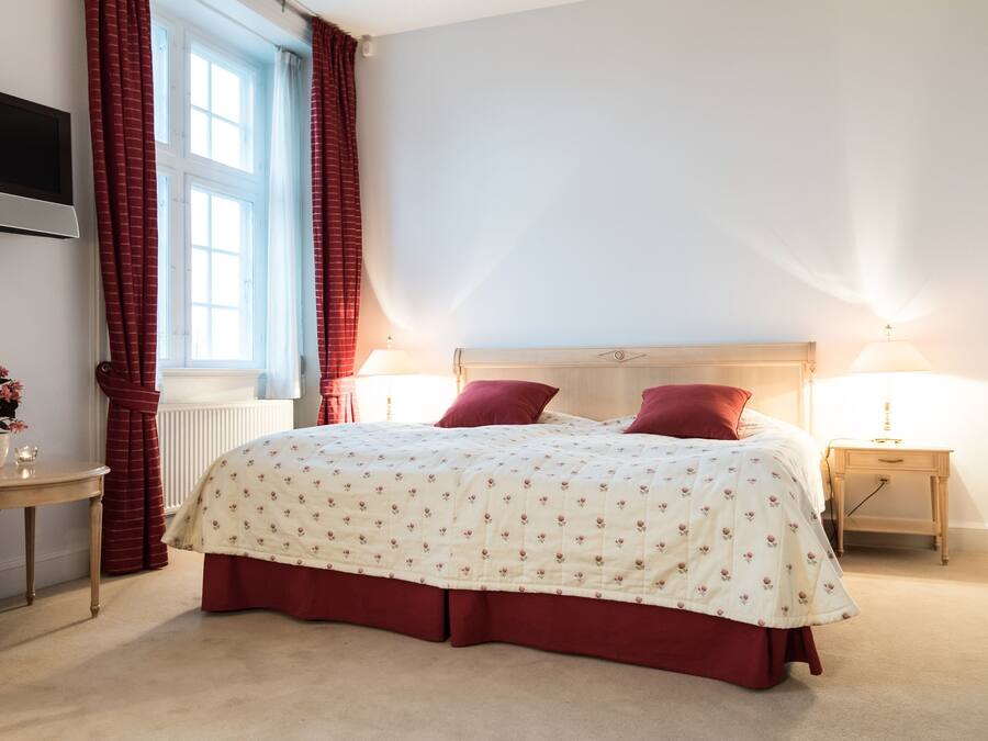 Quarto com cama, duas luminárias de cabeceira, televisão na parede, janela com cortinas e uma mesinha de centro com um vaso de flores.