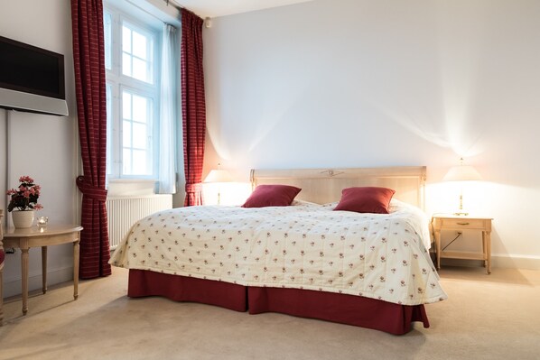 A bedroom with a bed, two bedside lamps, a TV mounted on the wall, a window with curtains, and a small table with a flower vase.