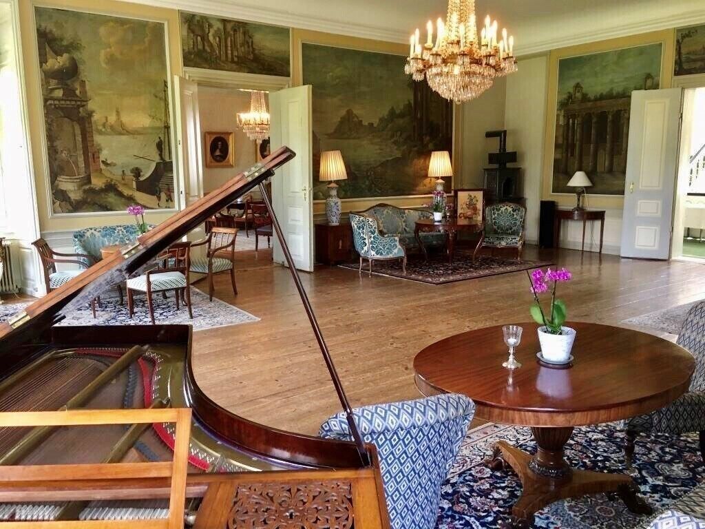 Una sala imponente con un piano de cola, una araña y una mesa redonda con un jarrón de flores.