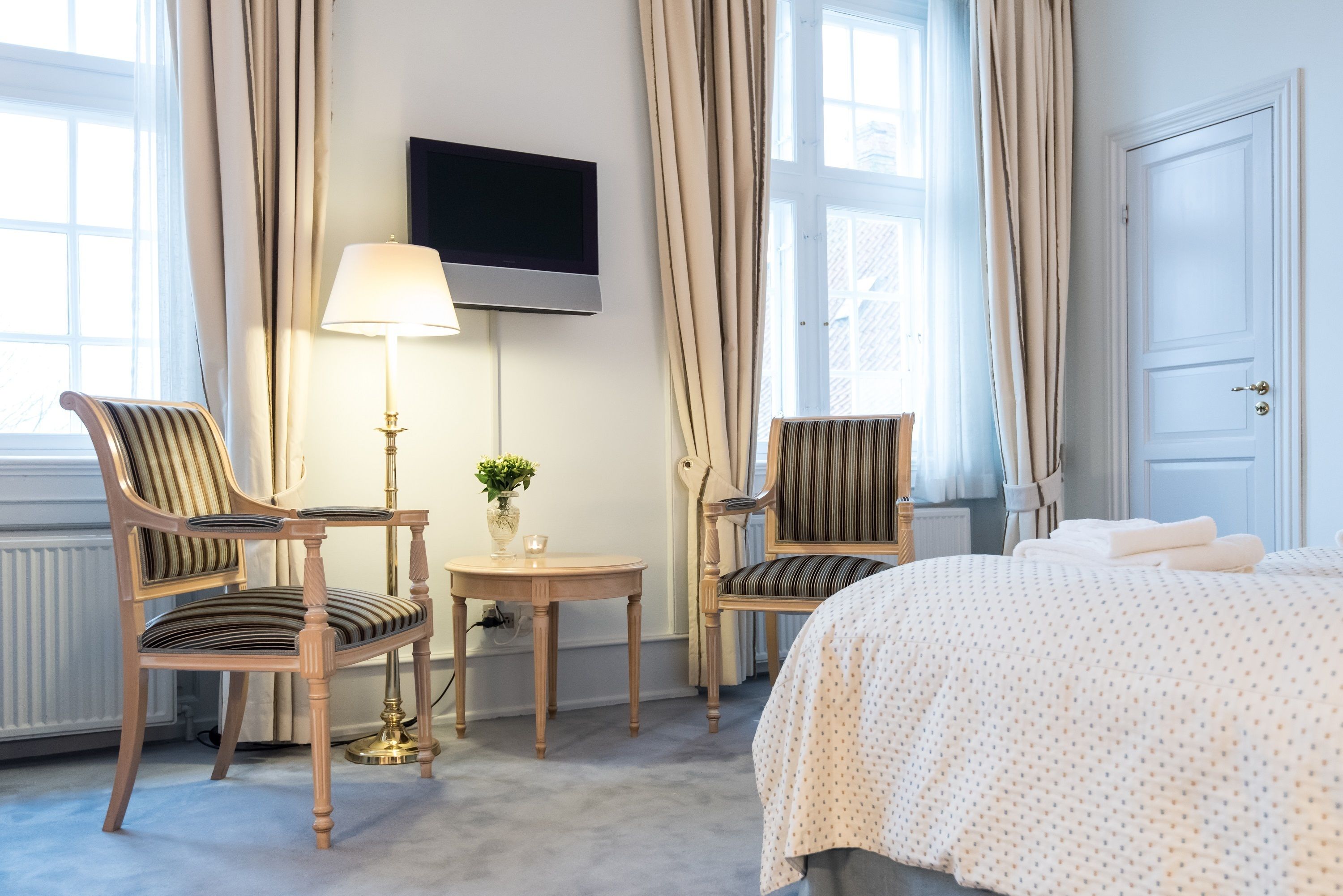 A hotel room with a bed, two chairs, a table with a lamp, a small plant, and a TV on the wall.
