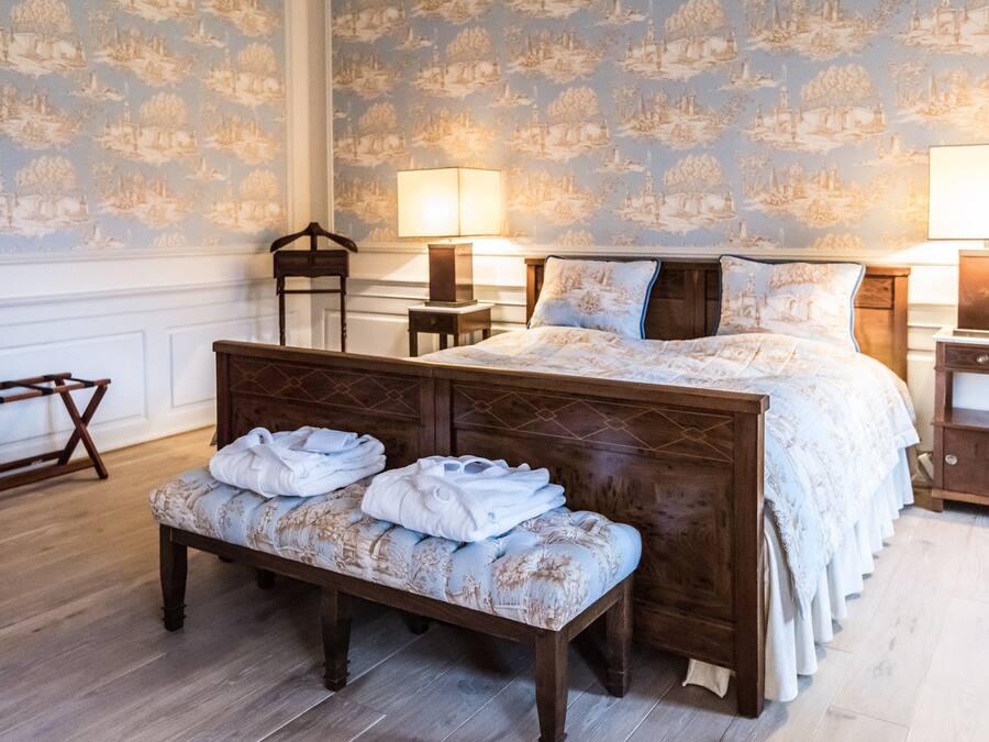 Un dormitorio ordenado con una cama de madera, dos mesitas de noche, un banco con toallas y un papel tapiz estampado.