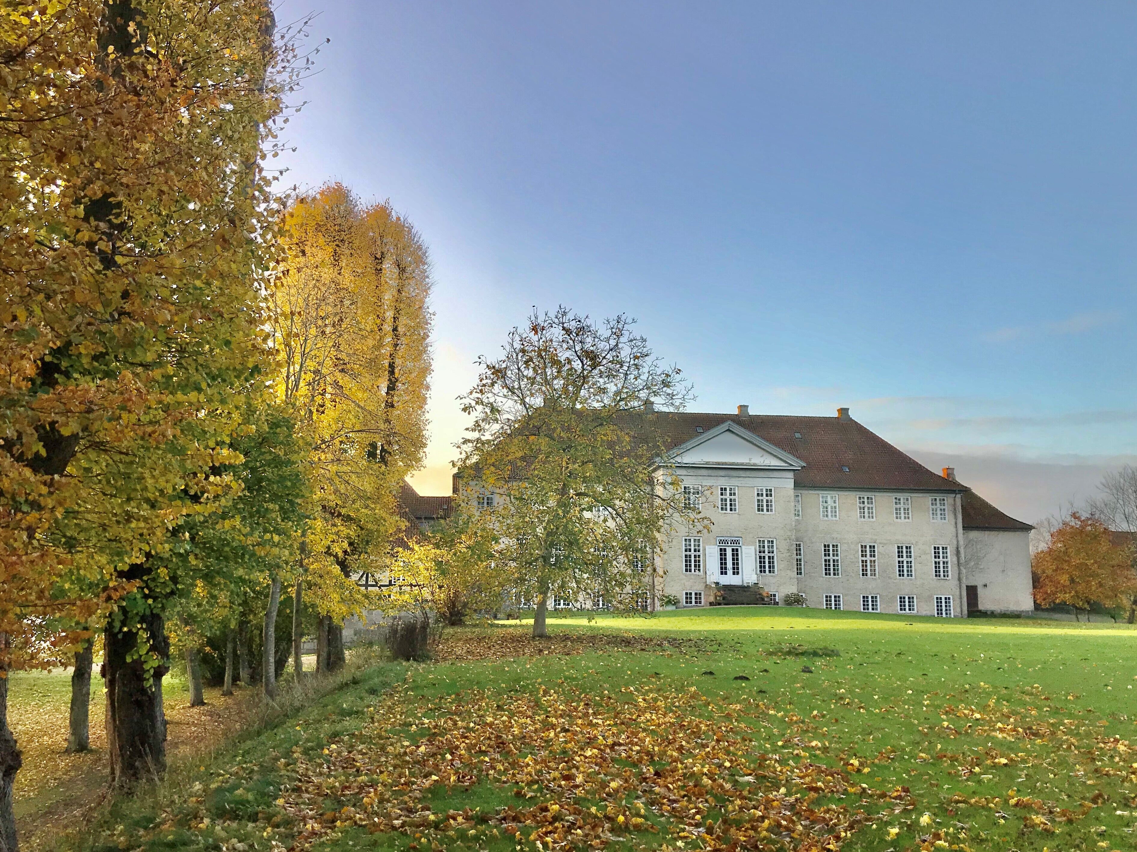 Un grand bâtiment historique à la façade symétrique, entouré d’un champ verdoyant et d’arbres aux feuilles d’automne.