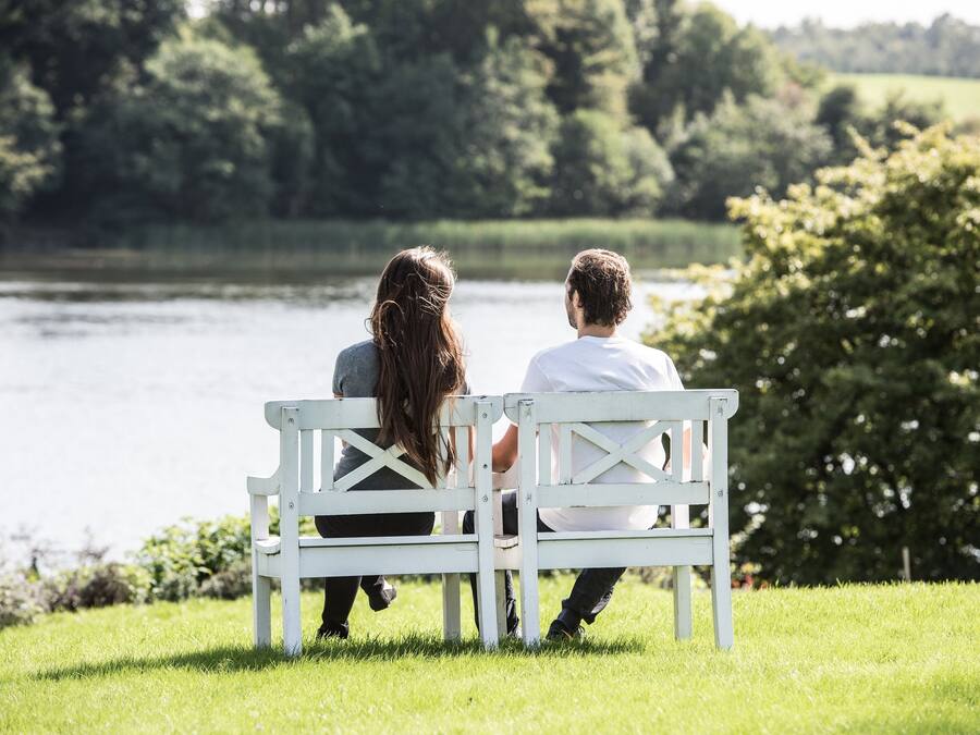 A white bench with a cross-back design in a grassy area with a lake and trees in the background.