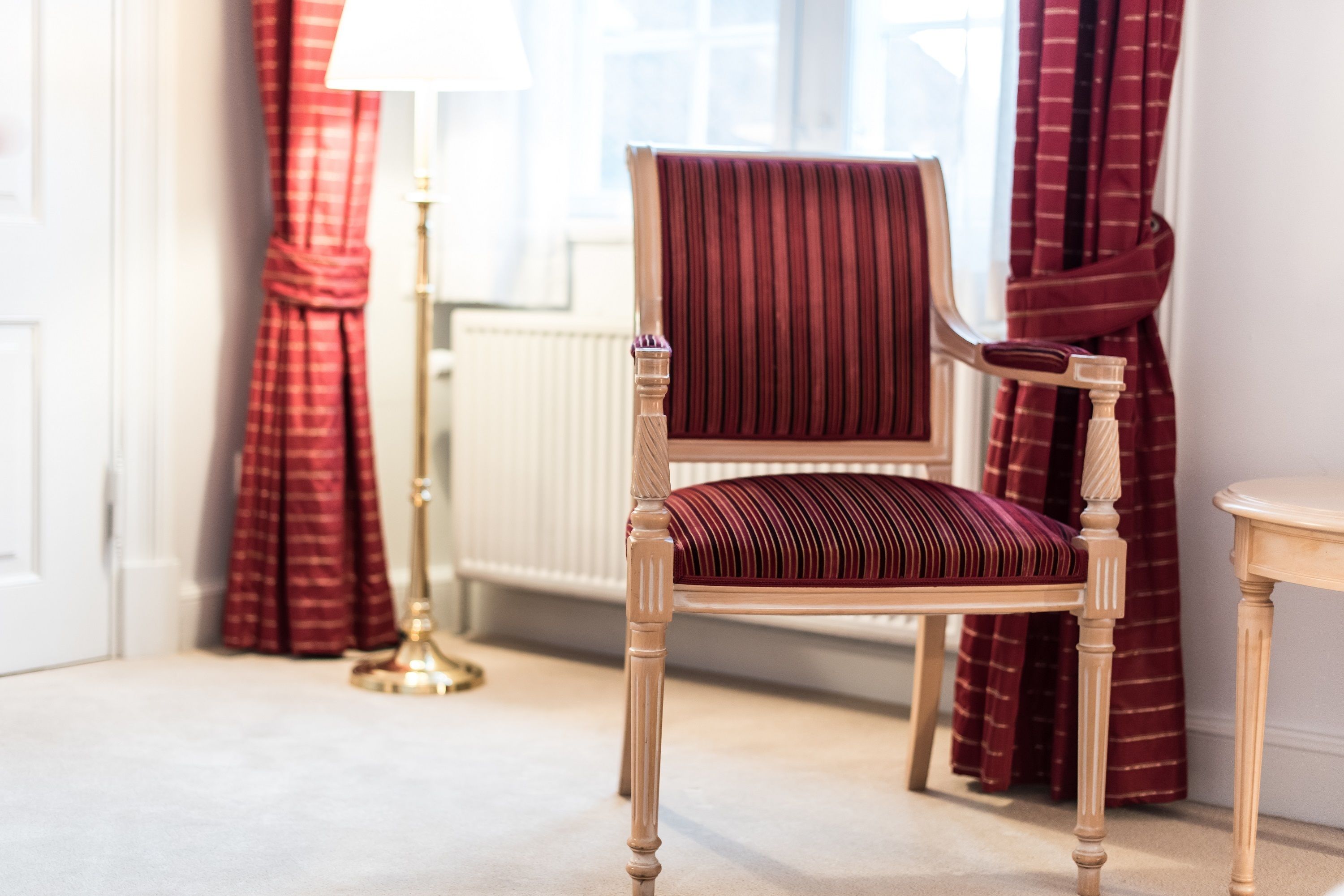 A wooden chair with a cushioned seat and backrest, positioned in a room with a radiator, curtains, and a lamp.
